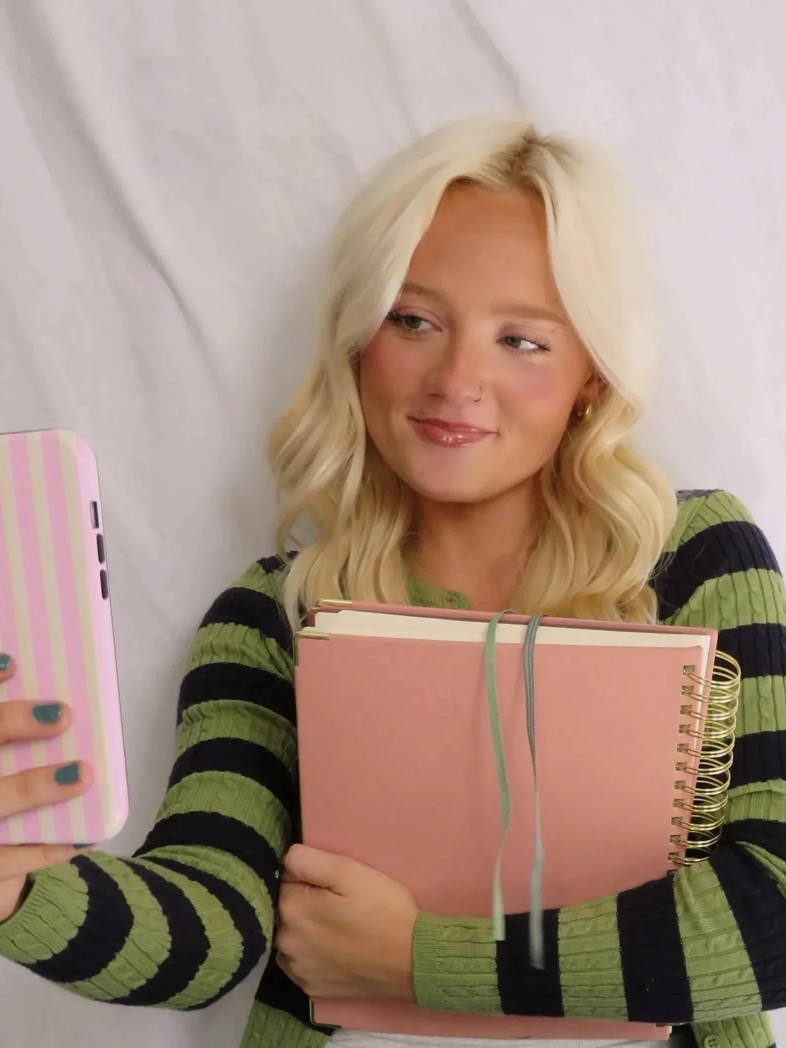 A young woman with blonde hair taking a selfie in front of a plain white wall, holding pink notebooks. She is wearing a green and black striped sweater and has makeup with glossy lips.