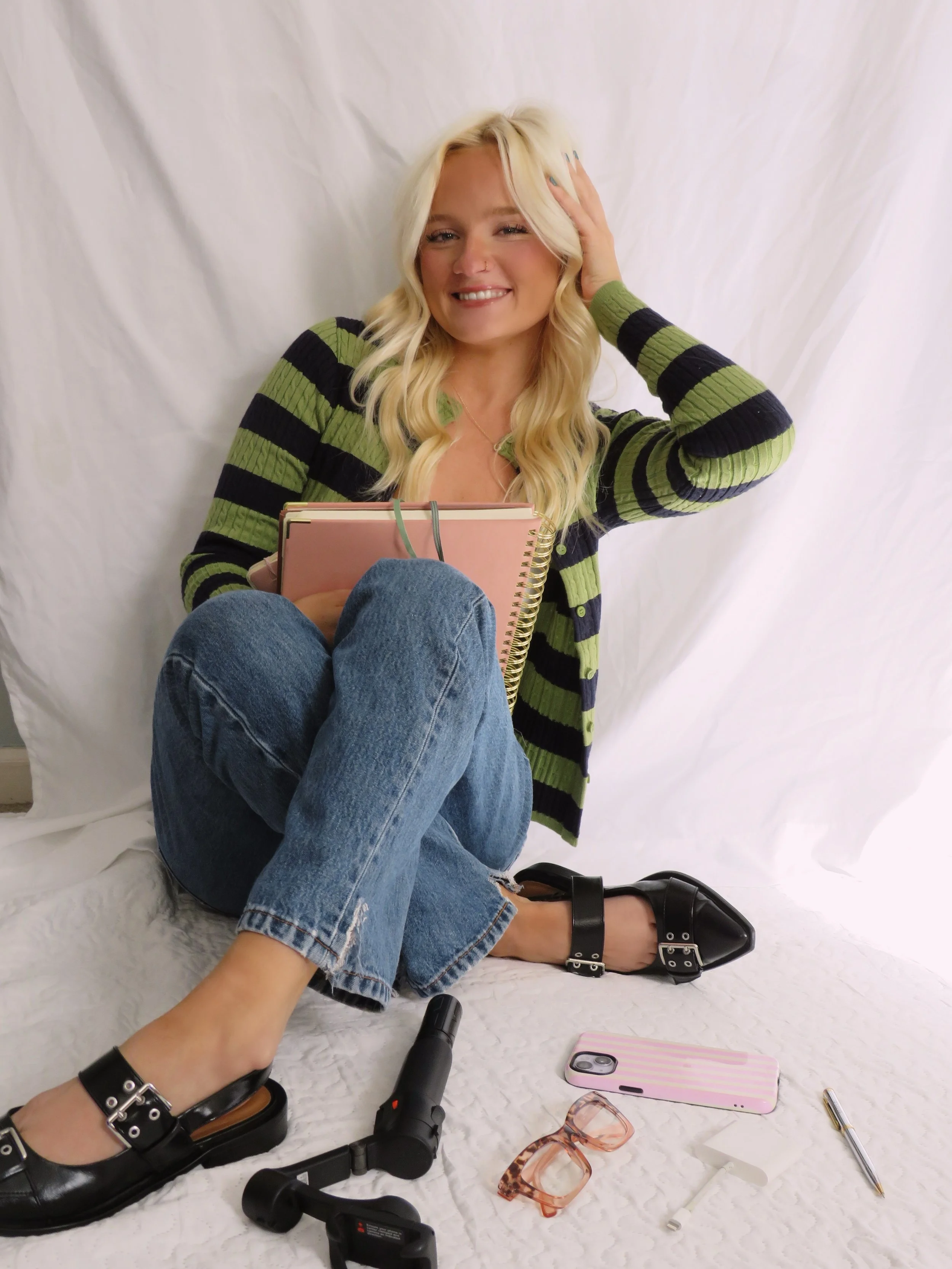 Young woman with blonde hair, wearing a striped green and black cardigan, sitting against a white backdrop with her eyes closed and smiling, holding pink notebooks. Items on the floor include a hairdryer, sunglasses, a pen, a phone with a pink striped case, and various makeup and accessories.