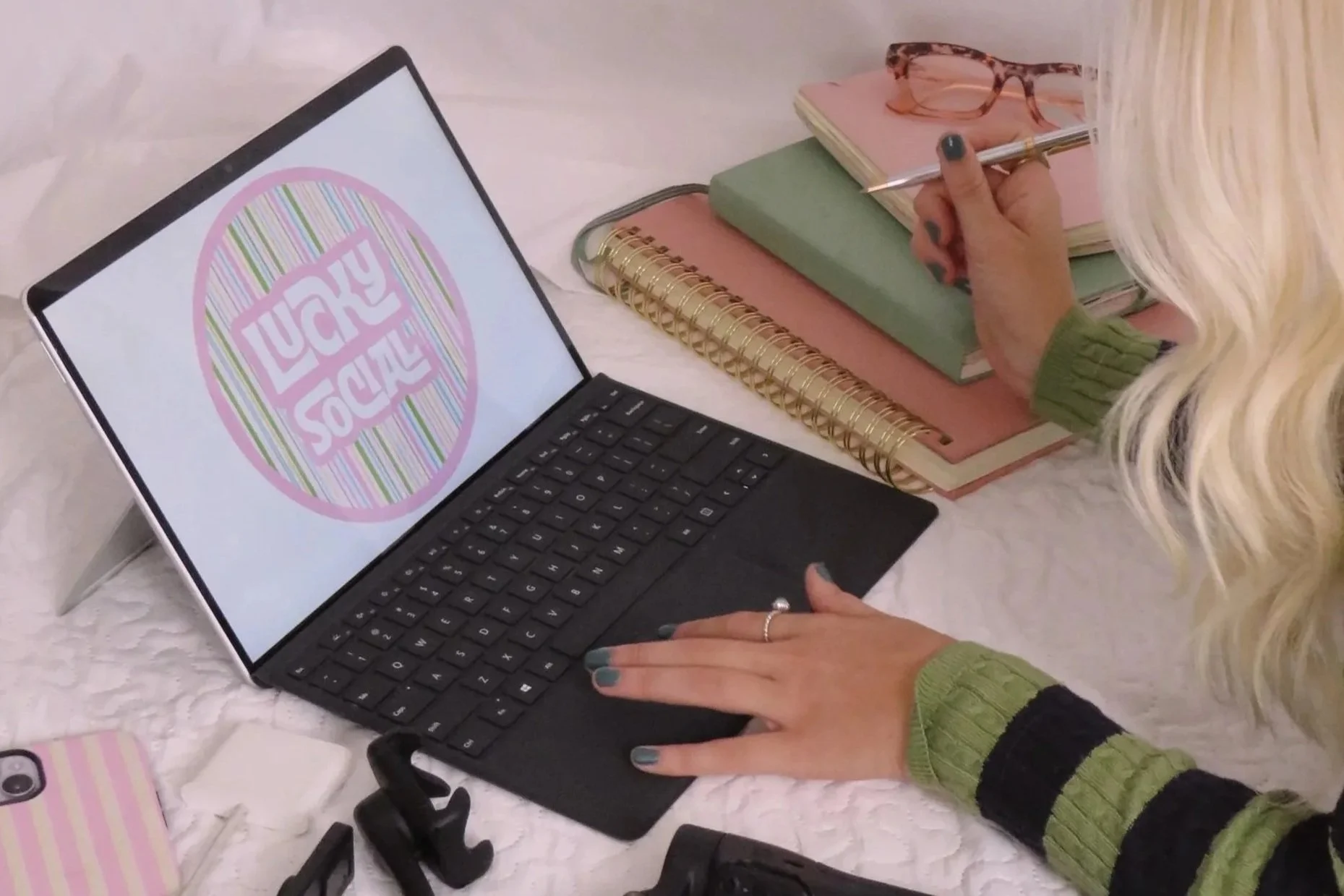 A person with blonde hair wearing a green and black striped sweater is sitting in front of a laptop on a white table. The laptop screen displays a colorful circle logo with the words "LUCKY SOCIAL". The person is holding a pen and writing on a stack of pastel-colored notebooks stacked on top of spiral-bound notebooks. On the table, there are additional notebooks, a pink and tortoiseshell glasses, a smartphone, and some black item straps.