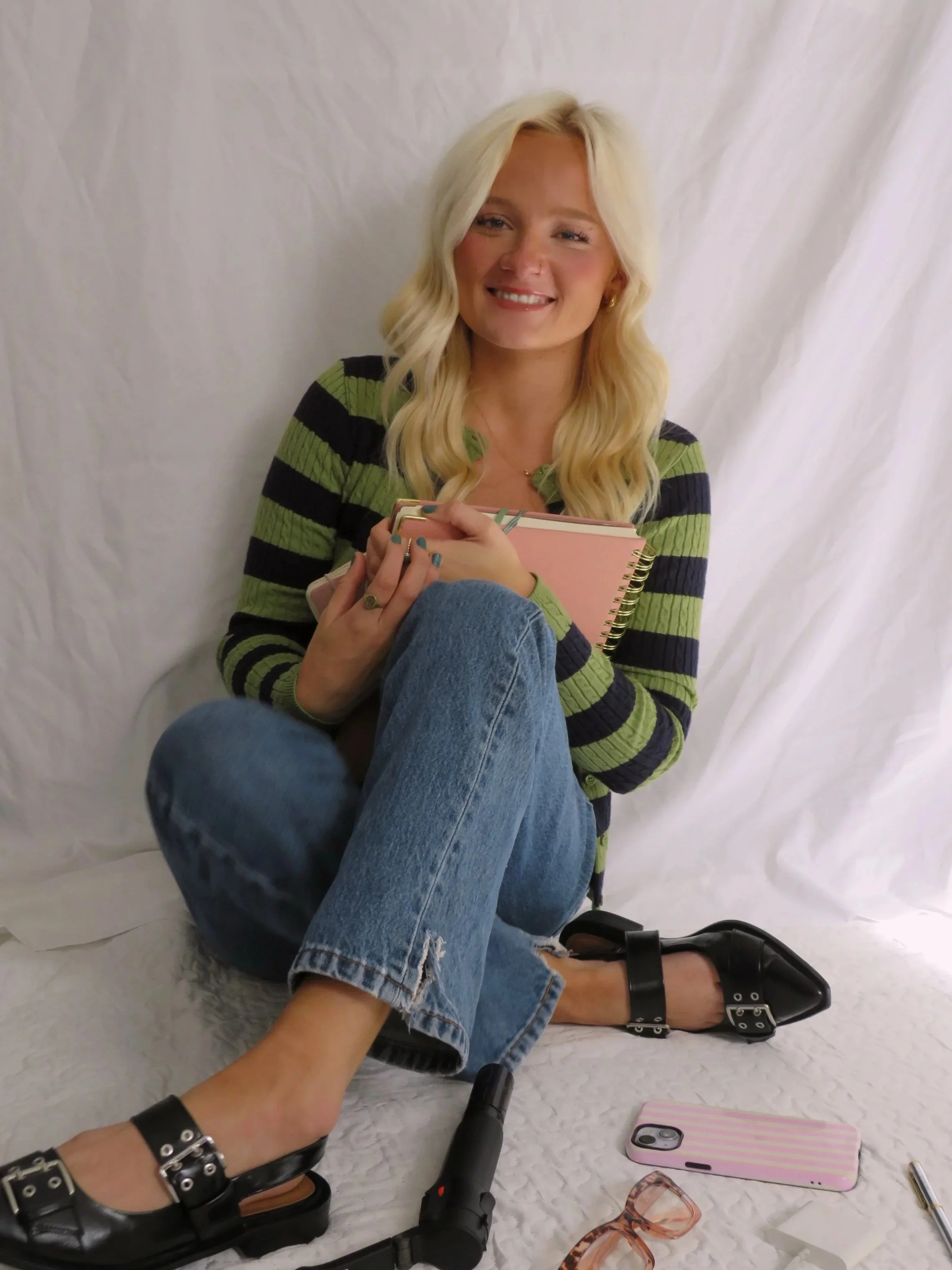 A woman with long blond wavy hair, wearing a green and black striped shirt, is sitting on a white fabric backdrop, smiling and holding a pink notebook. She is wearing black shoes with ankle straps, gold earrings, and a necklace. Several personal items are scattered around her, including a phone with a pink case, reading glasses, and a flashlight.