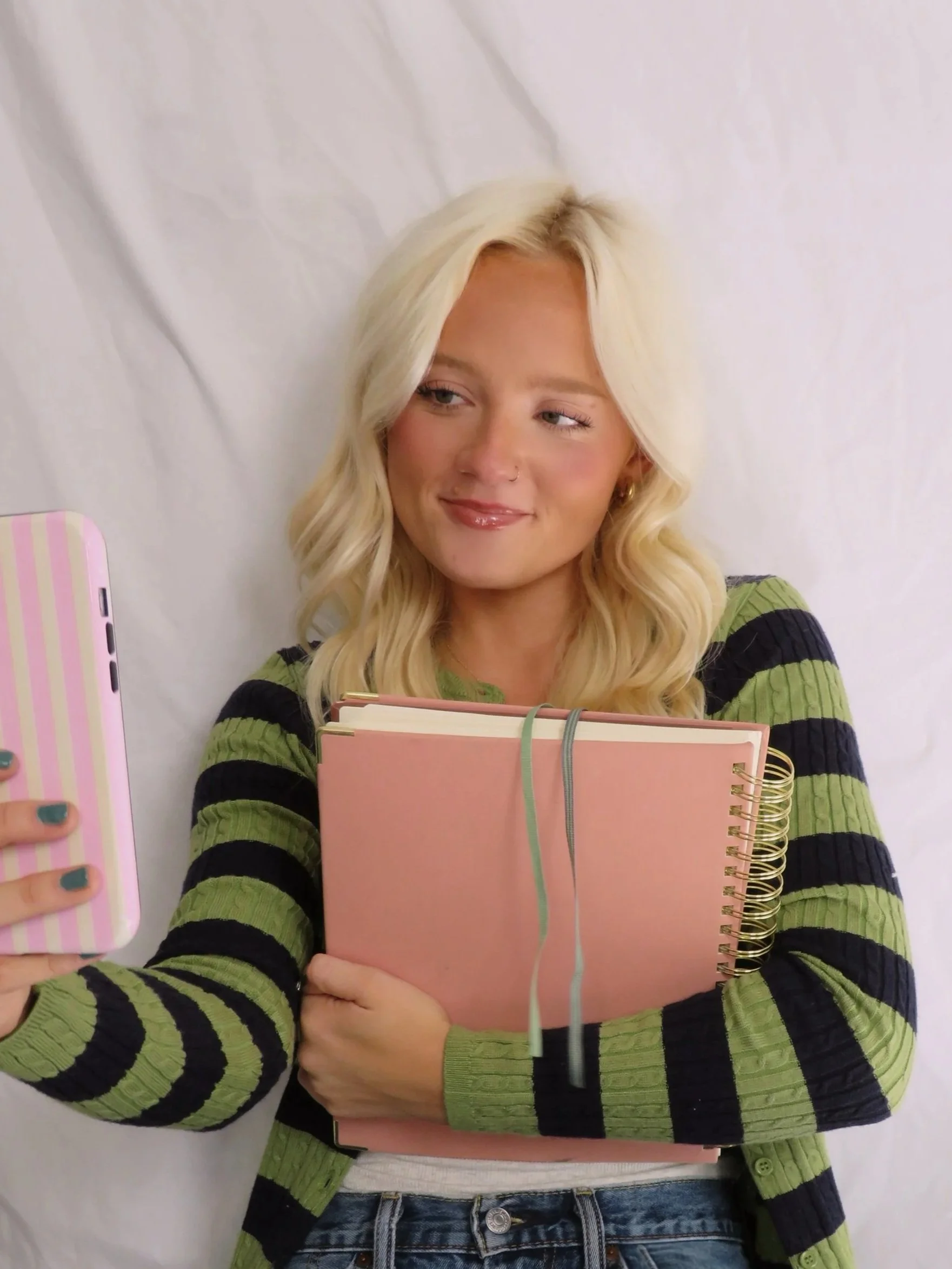 A woman with long blonde hair taking a selfie in front of a mirror, holding a pink notebook and wearing a black and green striped sweater.