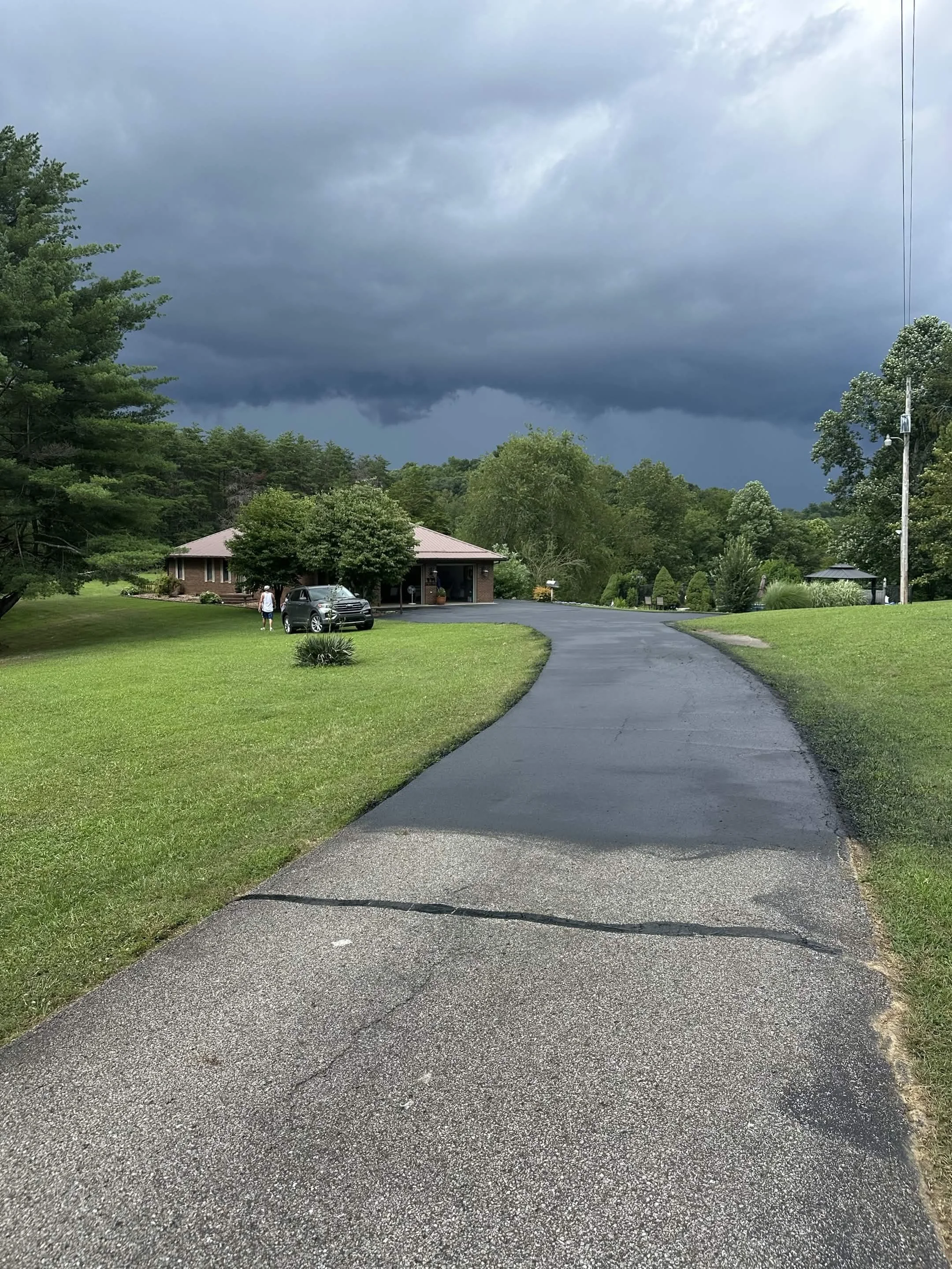 A driveway with dark asphalt leading to a house with a brick exterior. The sky is overcast with dark clouds, indicating potential stormy weather. There are trees and greenery surrounding the house.