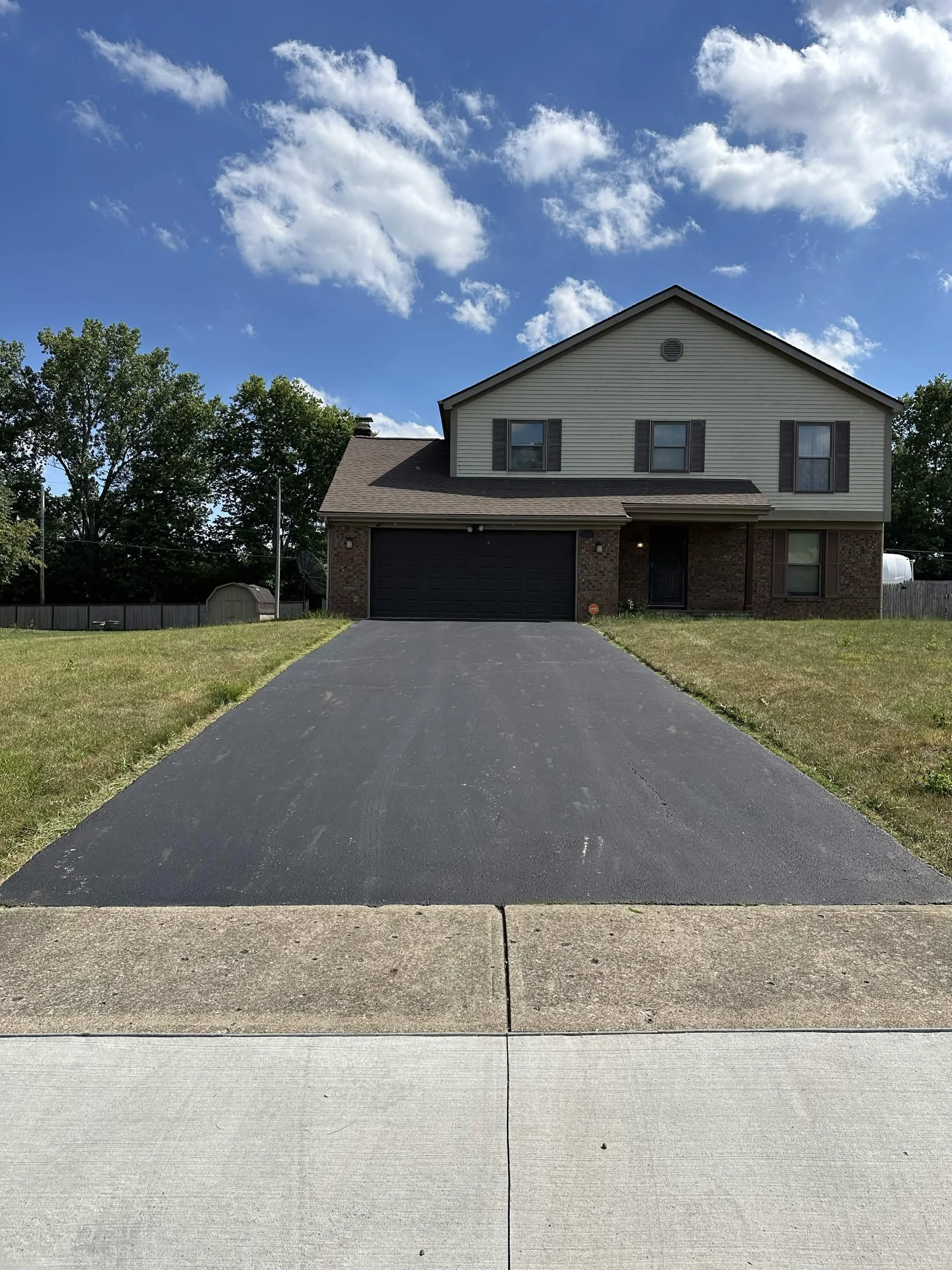 A two-story house with beige siding, black shutters, and a dark garage door, surrounded by a lawn, with trees and a blue sky with white clouds in the background.