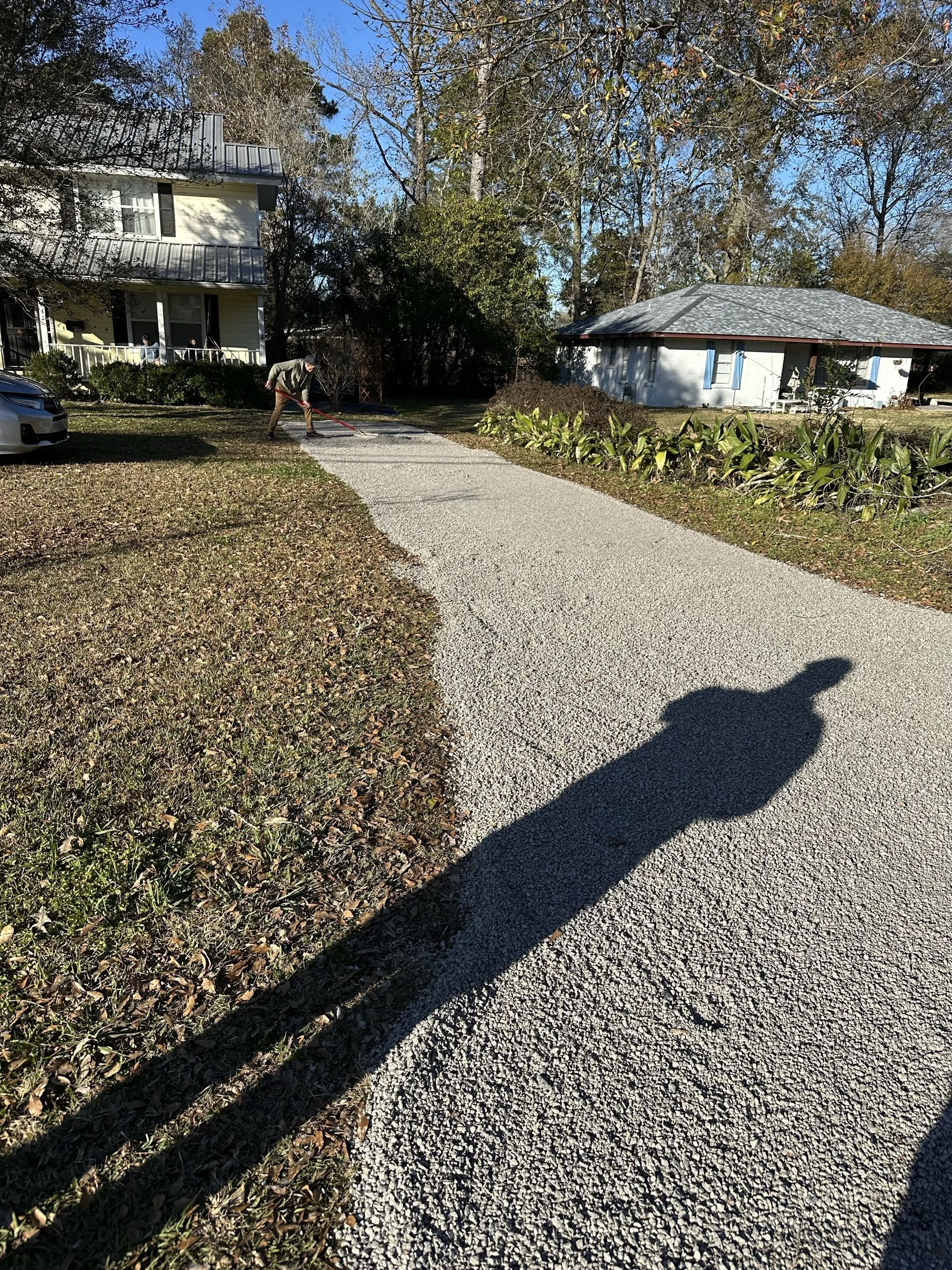 Person spreading gravel on a driveway in front of a house with trees and shrubs around.