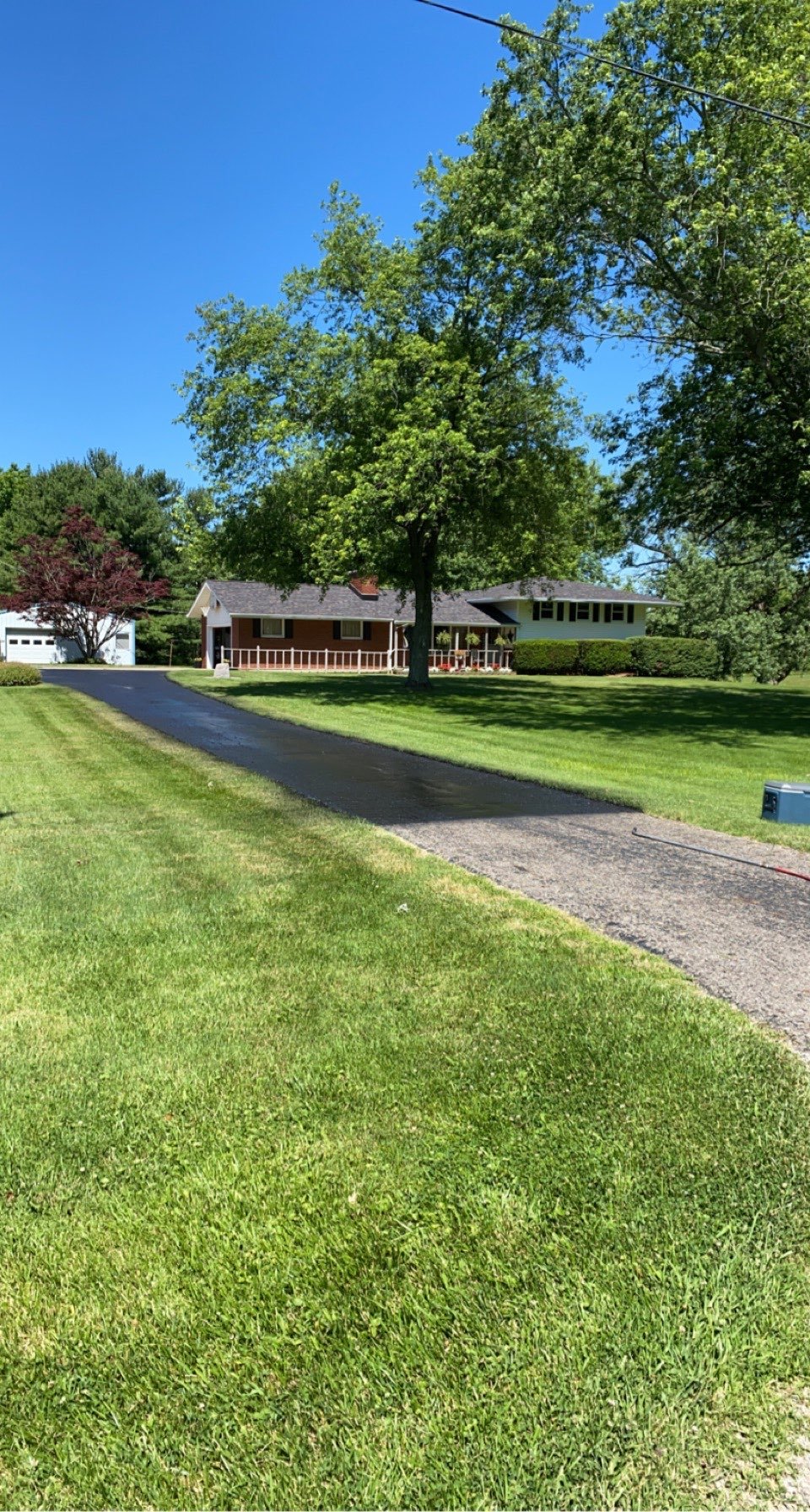 A residential house with a driveway, large trees, a well-maintained lawn, and a small shed or garage in the background on a clear, sunny day.
