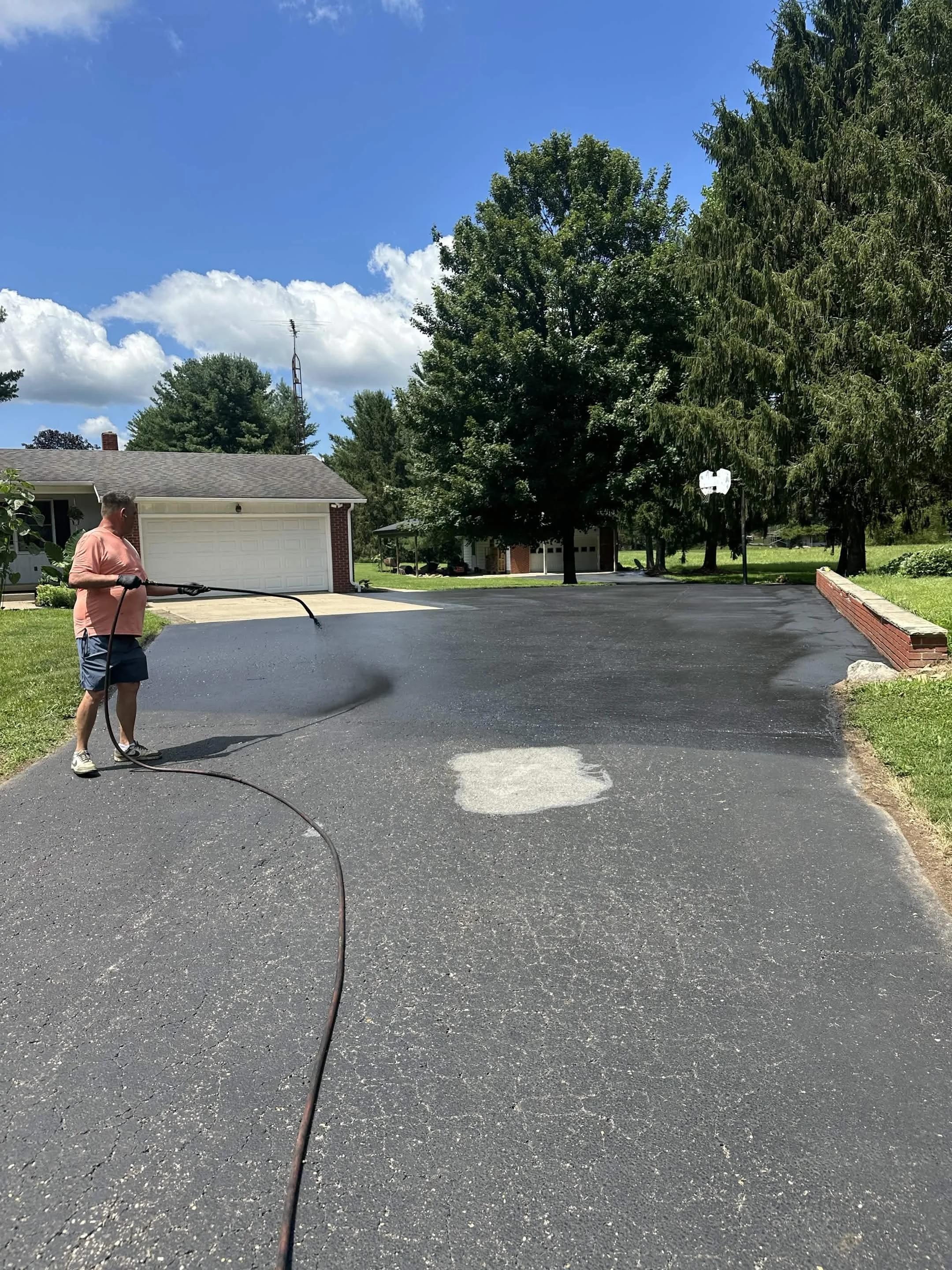 Man watering freshly laid asphalt on a driveway in front of a house during the daytime, with trees and a blue sky in the background.