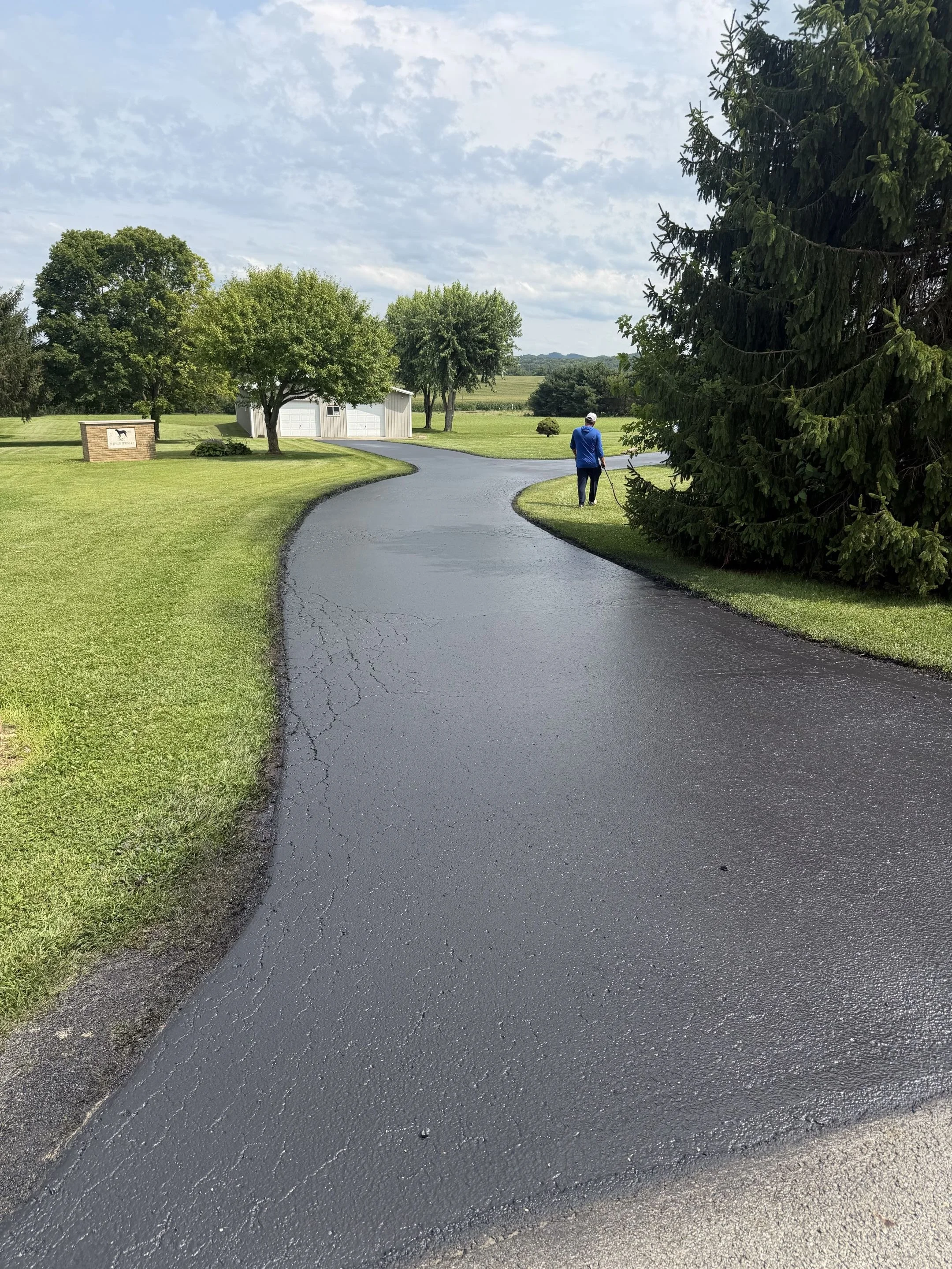 A person in a blue shirt, dark pants, and white cap sweeping a freshly paved black asphalt driveway with a broom.