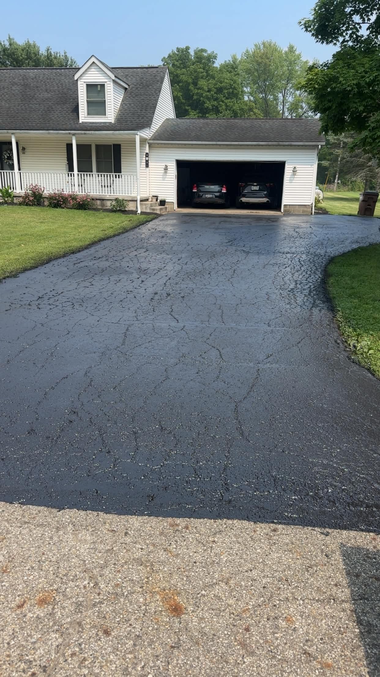 A house with a driveway leading to a garage, partly paved with asphalt and partly concrete, with green lawn and trees in the background.