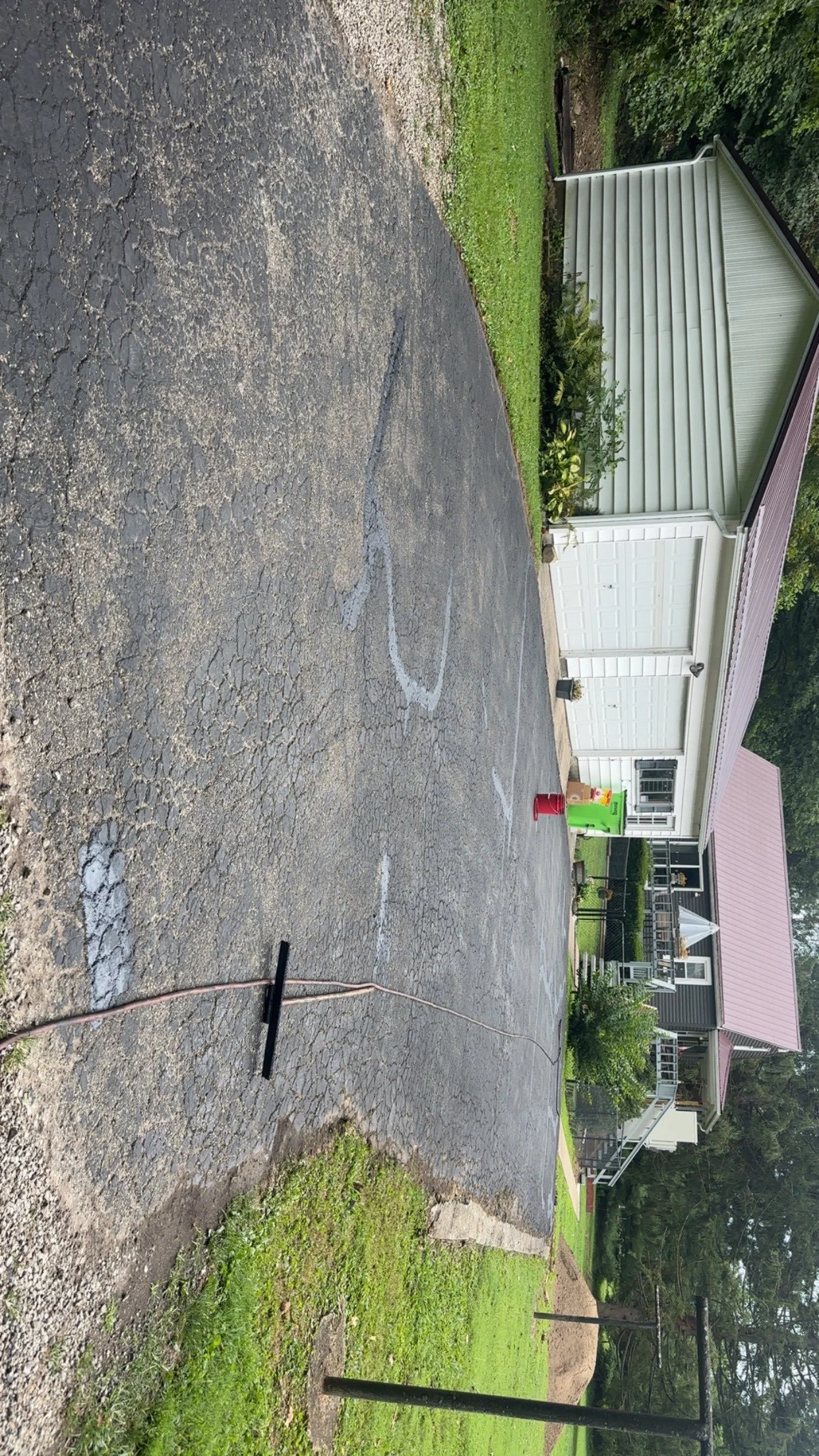 A residential driveway with a new layer of asphalt, marked with white lines for parking, adjacent to a house with white siding and greenery around.