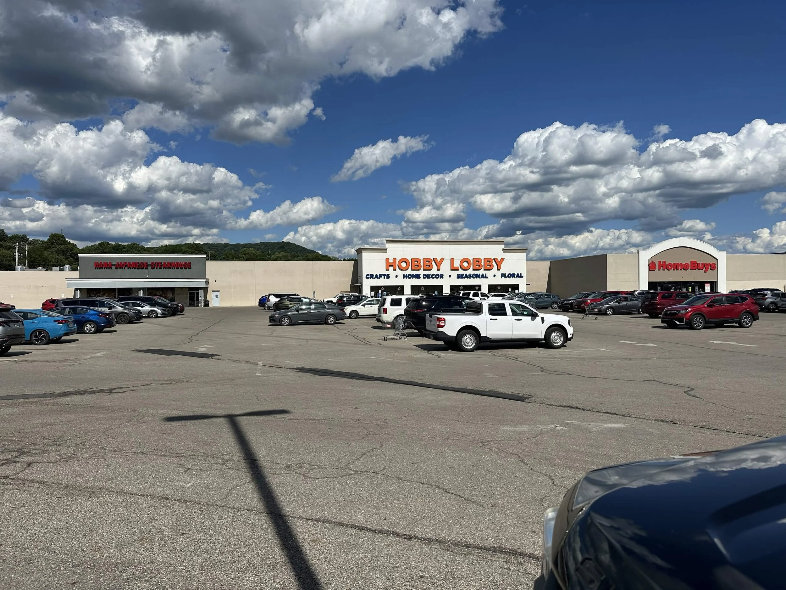 A parking lot in front of a shopping center with a sign for Hobby Lobby and other stores, filled with various cars under a partly cloudy sky.