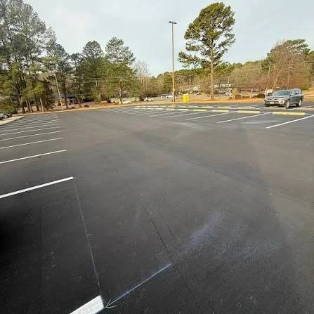Empty parking lot with a few cars in the background and trees beyond, under a clear sky.