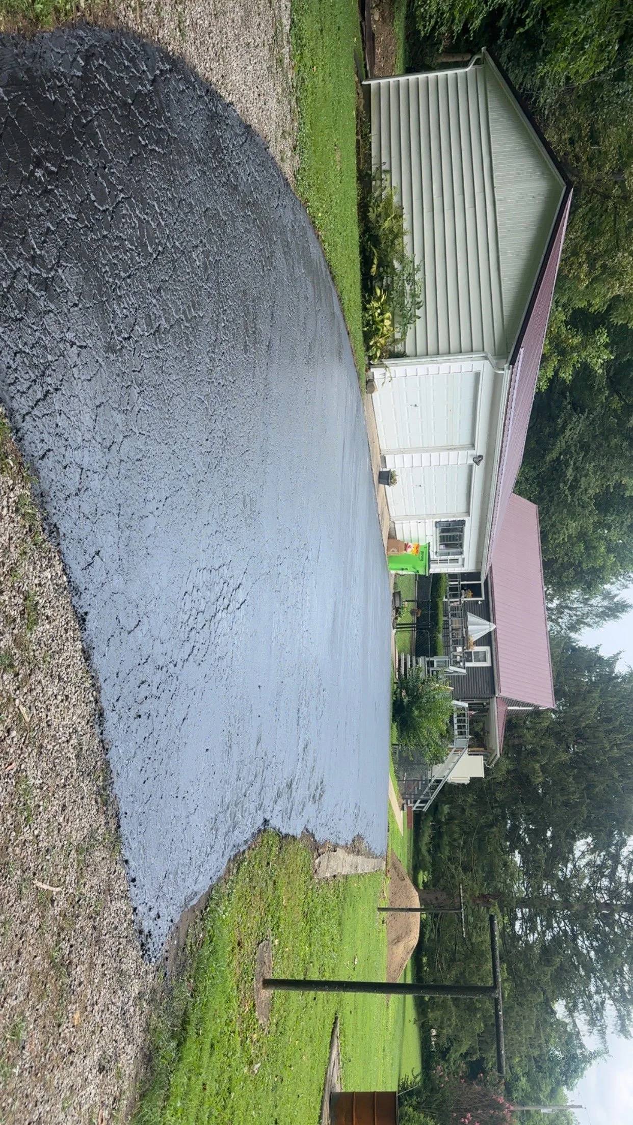 A freshly paved driveway leading to a house with a red roof. The house has white siding and a porch with white railings. There are trees and greenery surrounding the house, and a lawn with a few garden plants.