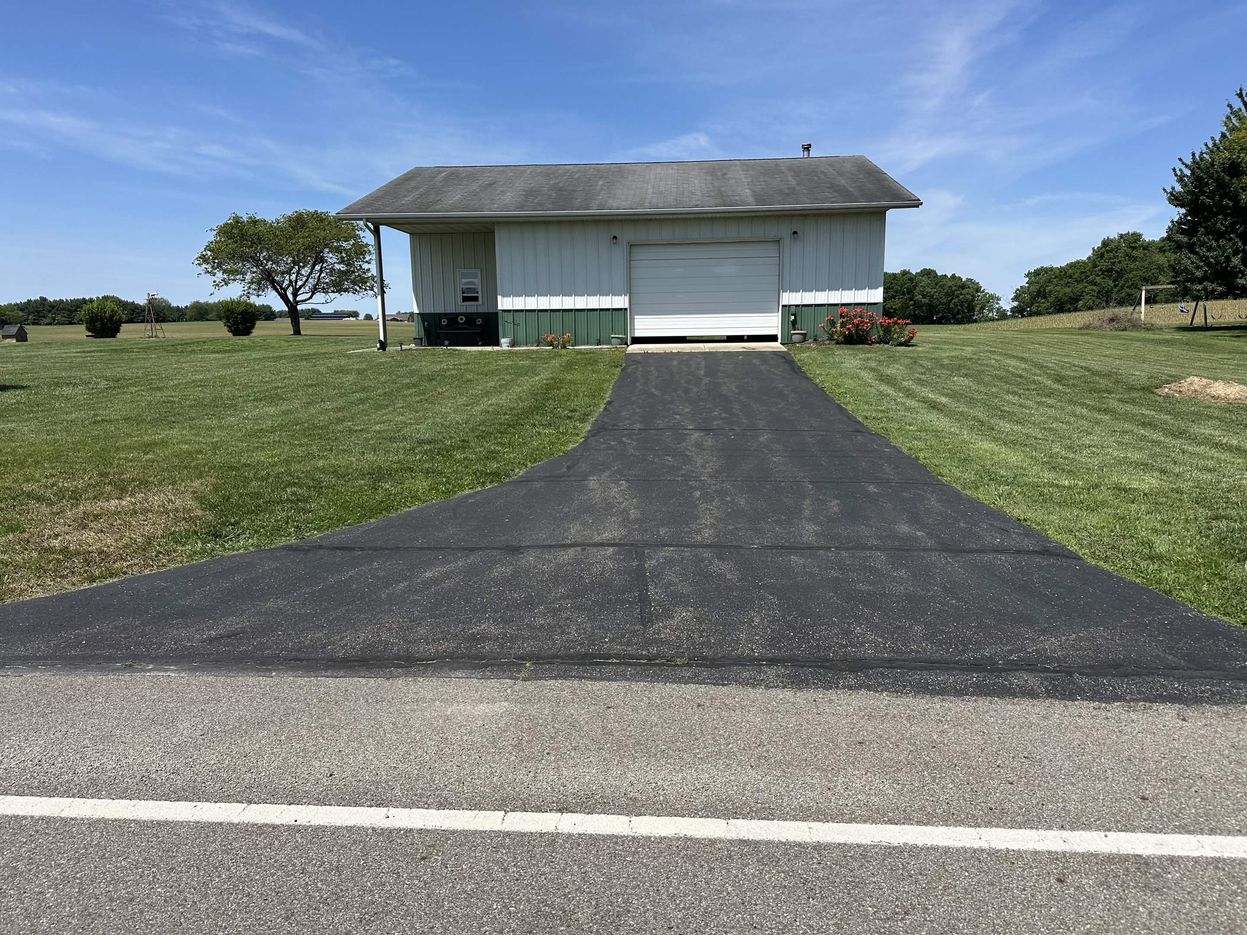 A rural scene with a garage, a driveway, a grassy field, trees, and a blue sky with some clouds.