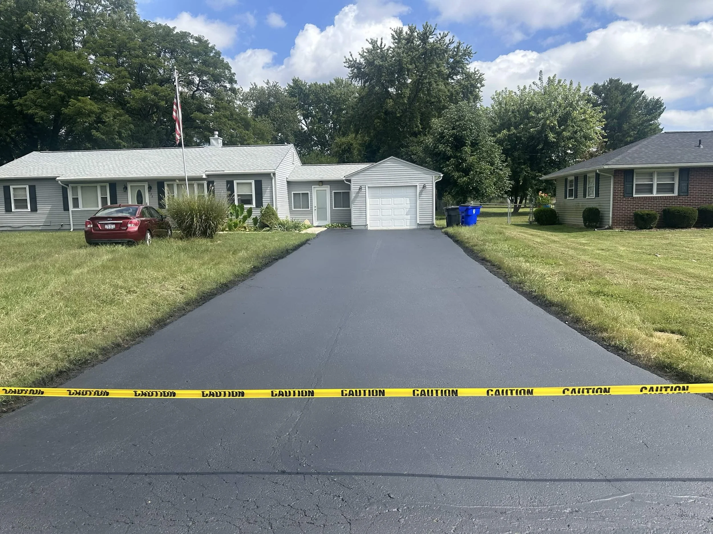 Residential driveway with caution tape across, garage and house visible, trees in background, blue sky and clouds overhead.