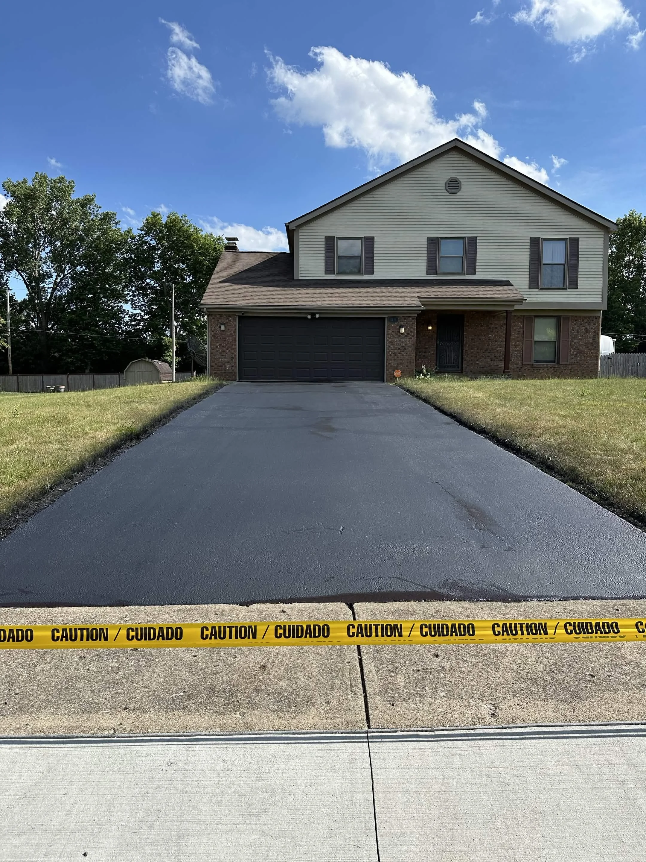 Photo of a residential house with a driveway, surrounded by grass and trees, and a yellow caution tape across the driveway with the words 'CAUTION / CUIDADO'.