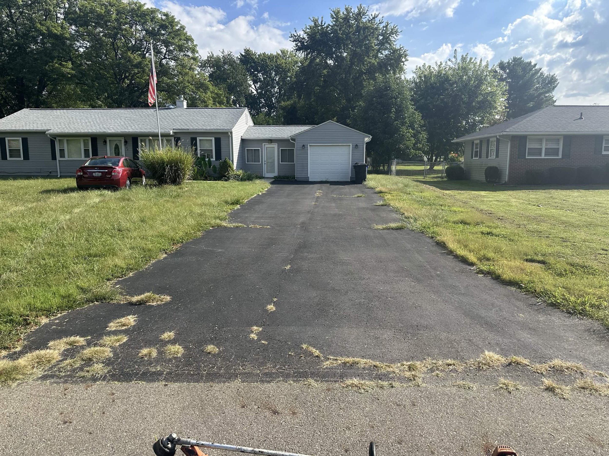 A driveway leading to a house with gray siding, a garage, and an American flag. There are trees in the background and the sky is partly cloudy.