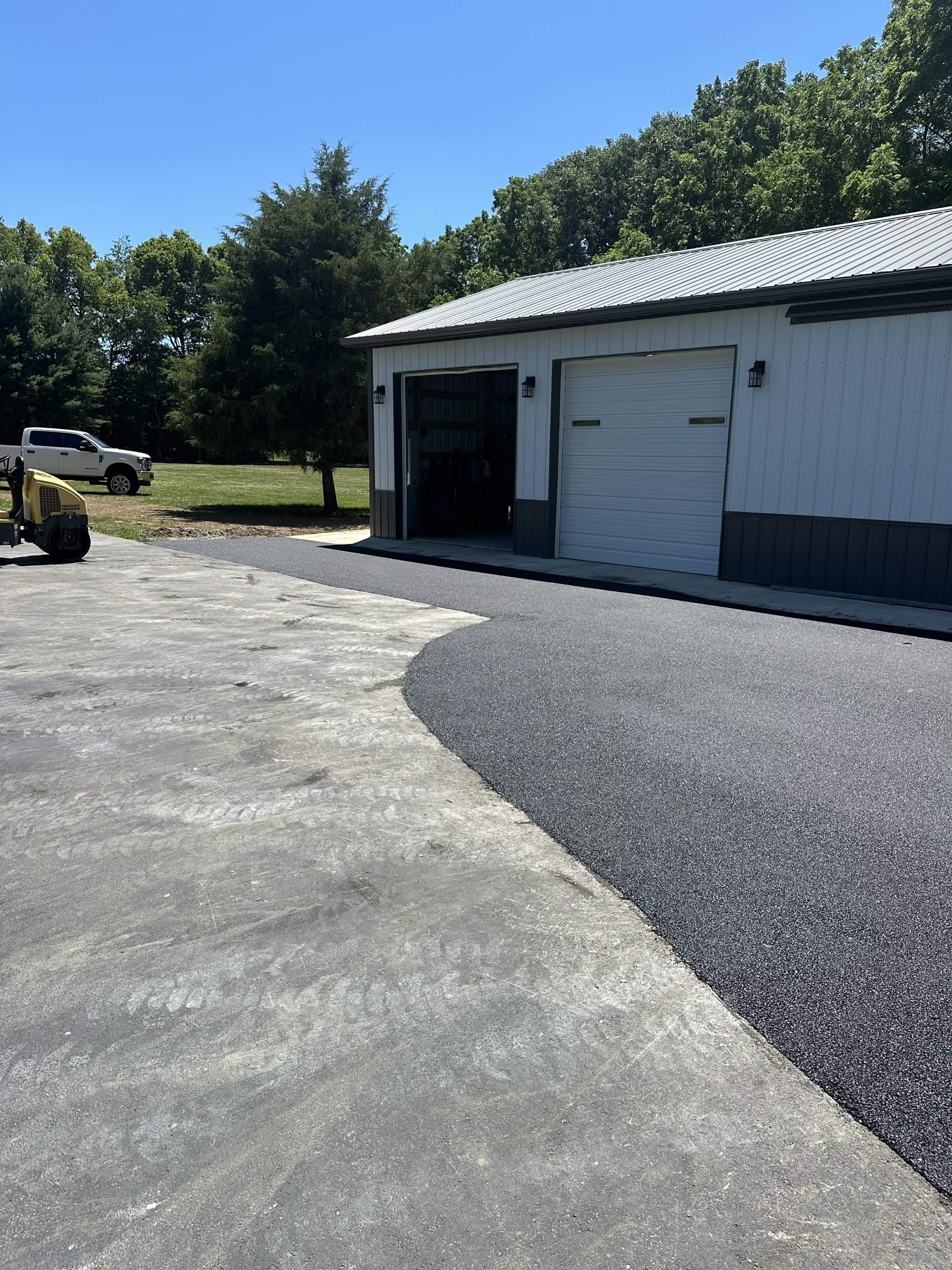 A newly paved driveway leading to a garage building with white metal siding, a rolled-up garage door, on a sunny day with a clear blue sky. A white truck and a yellow construction machine are parked nearby, with green trees in the background.