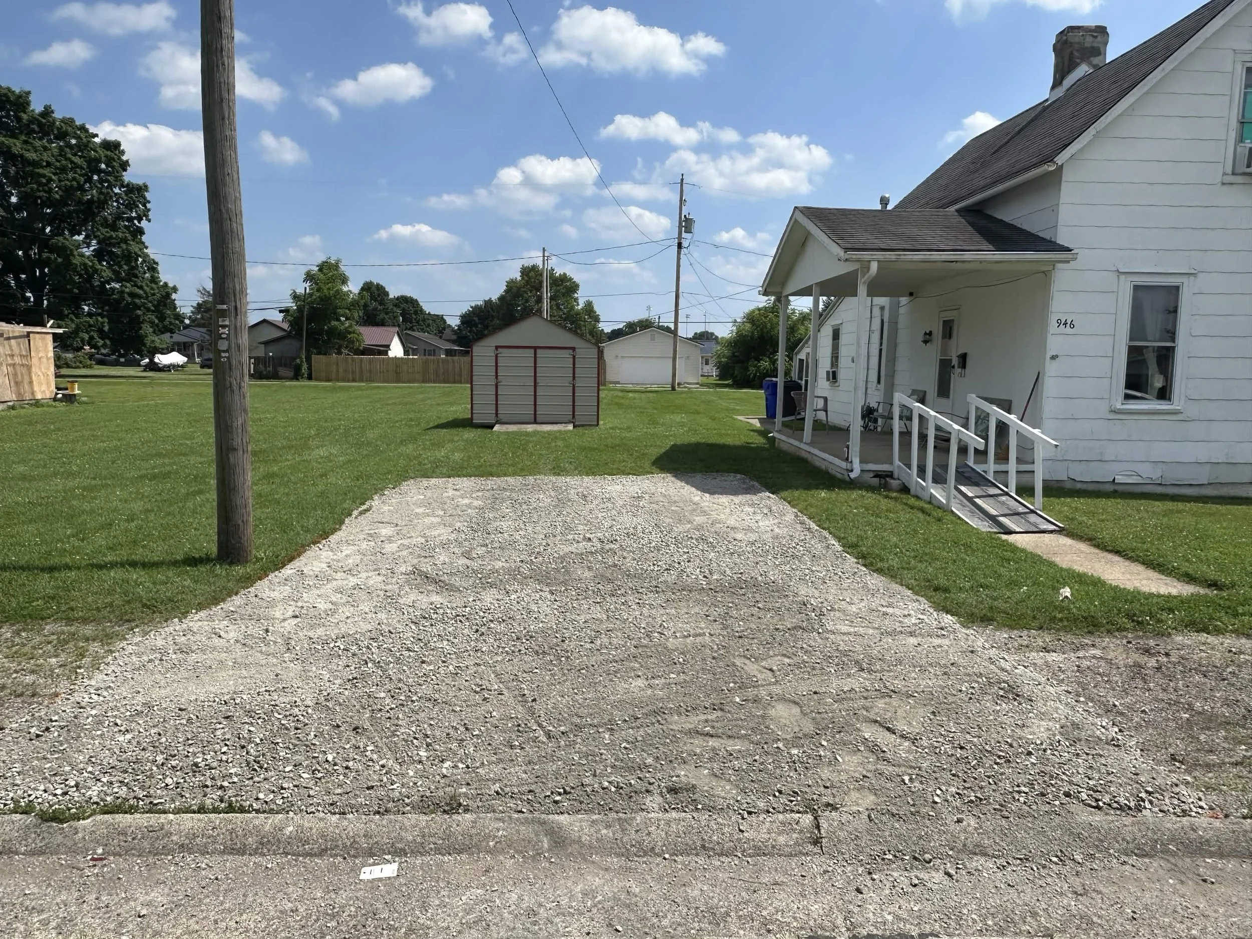 A gravel driveway leading to a white house with a porch, surrounded by green grass. There is a small shed in the yard, and power lines with poles. The sky is clear with a few clouds.