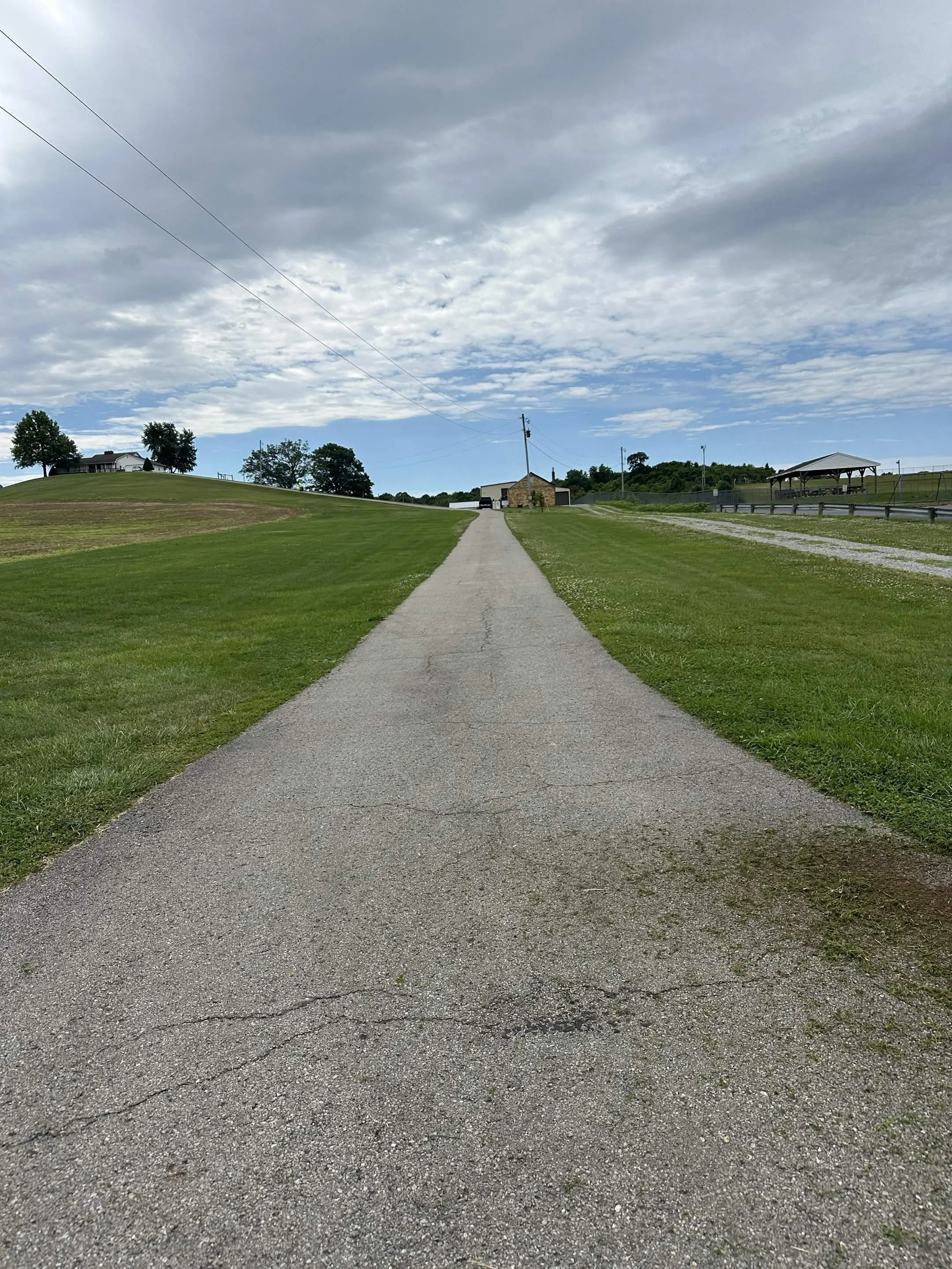 A paved sidewalk running through a grassy area towards a small building and a covered pavilion under a cloudy sky.