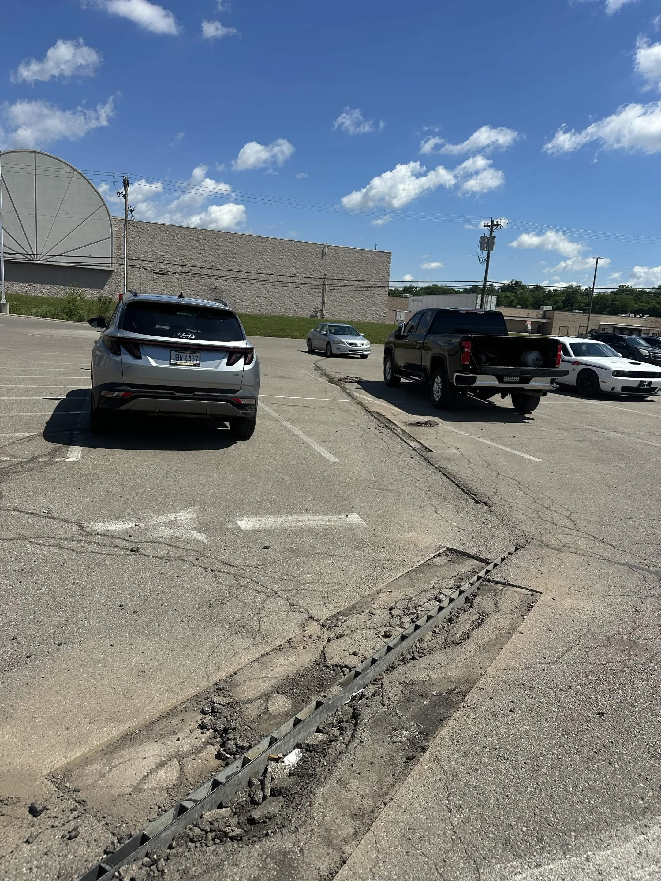 A parking lot with several cars parked and a large building in the background under a partly cloudy blue sky. There is a visible damaged section of the pavement with a metal drainage grate and cracked asphalt.