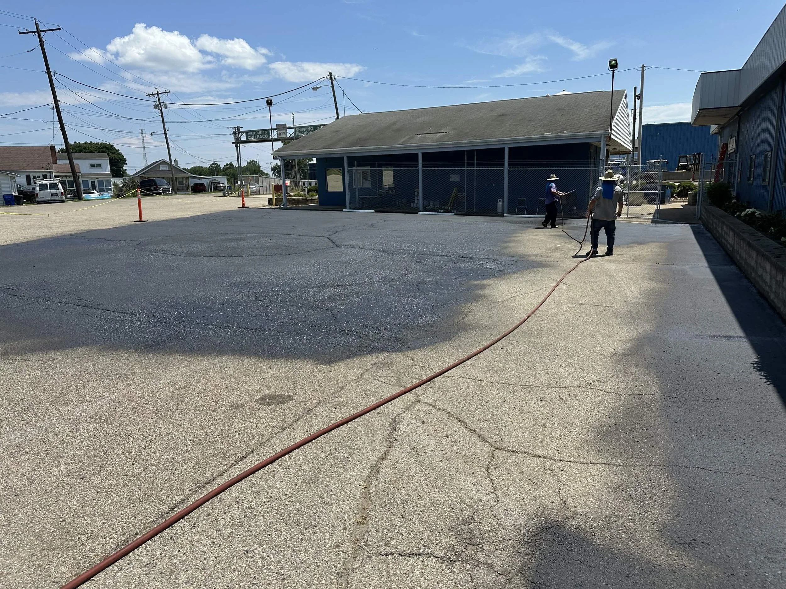 Workers power wash a parking lot surface near a blue building, with utility poles and residential houses in the background under a clear blue sky.