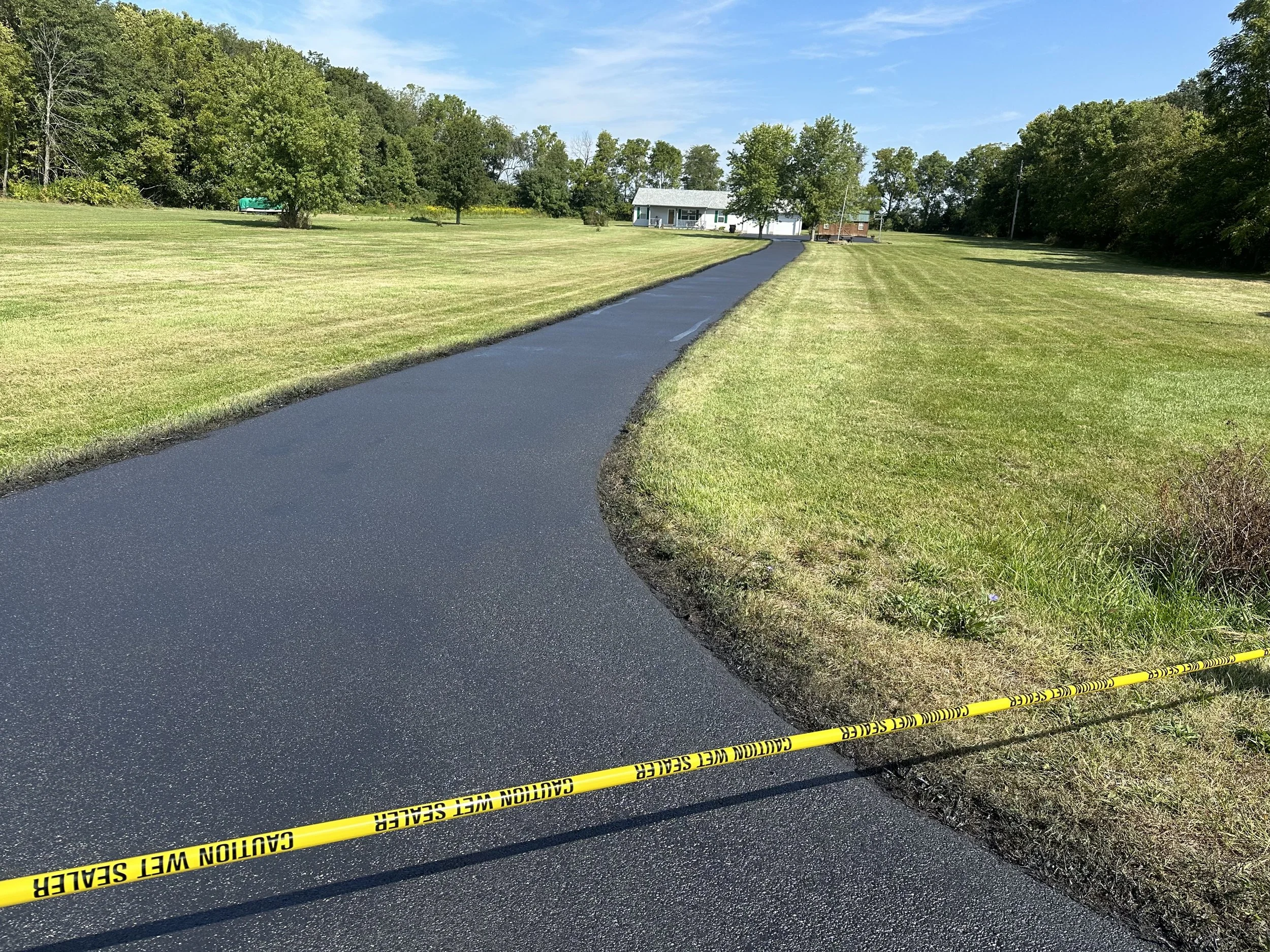 A newly paved black asphalt driveway leading to a white house with a gray roof, surrounded by green grass and trees. A caution tape reading 'Caution Wet Seal' is placed at the edge of the driveway.