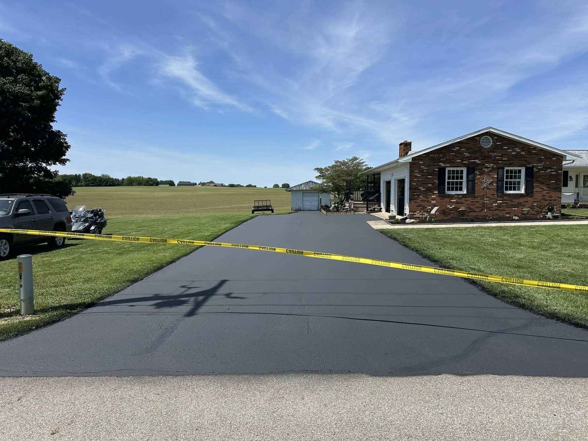 A paved driveway leading up to a house with brick and white siding exterior, situated in a rural area with open fields and a blue sky with some clouds.