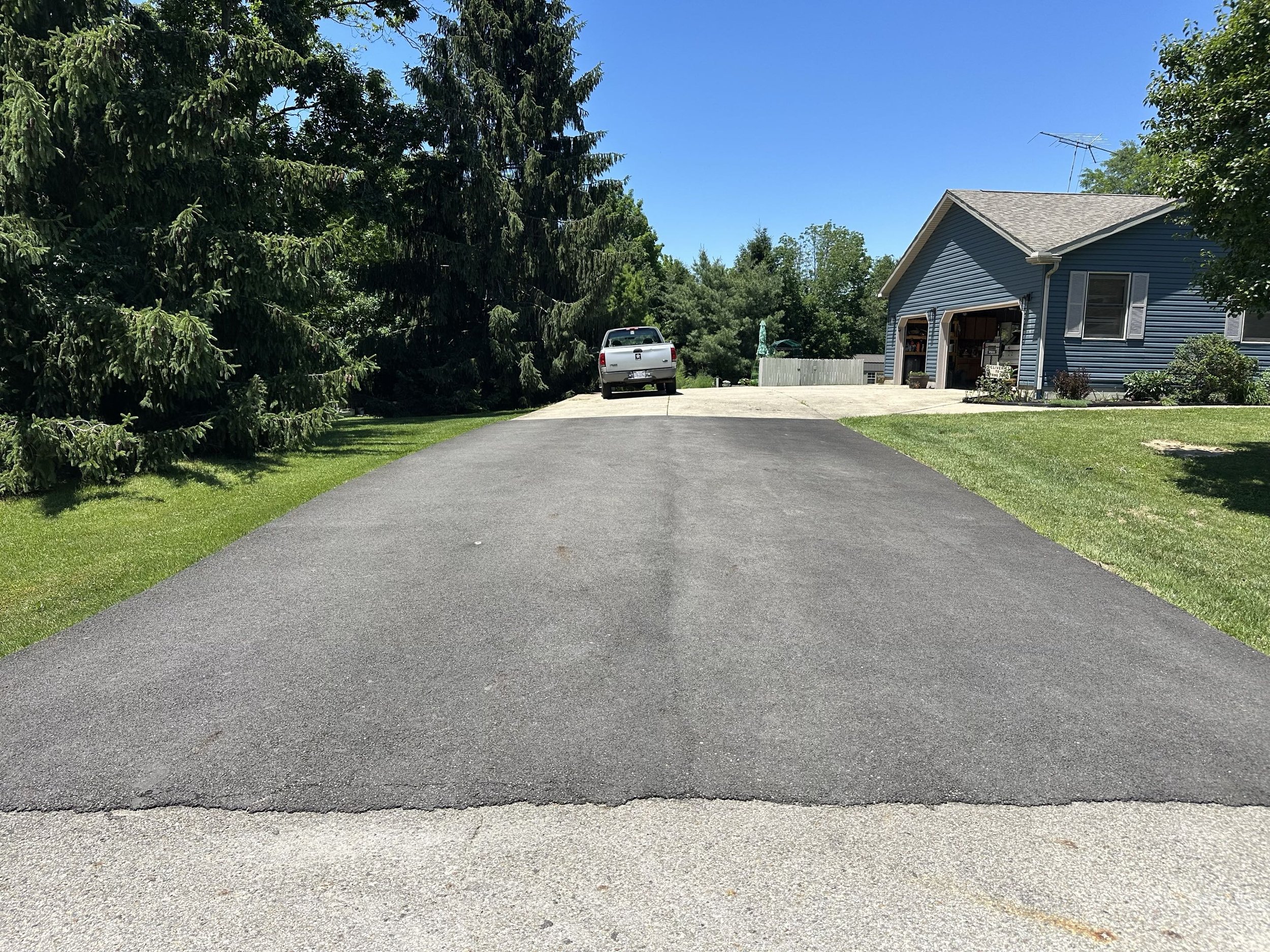 A driveway with a blacktop surface leading to a blue house with white trim and an open garage door. A white pickup truck is parked on the concrete section of the driveway near the house. There are trees and shrubs surrounding the house, with a clear 
