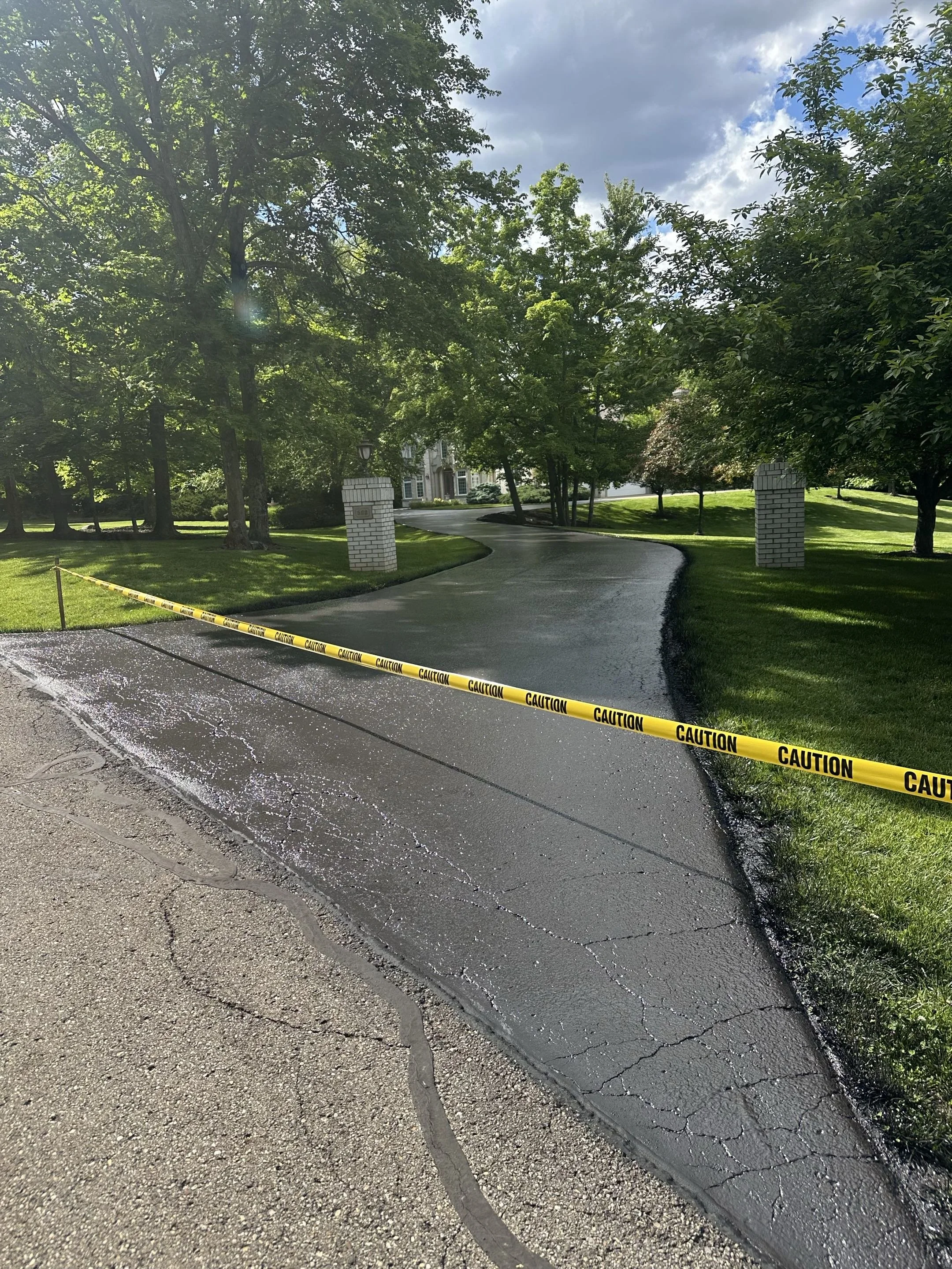 A paved driveway in a residential neighborhood with caution tape and flooding on part of the pavement, surrounded by green grass and trees, with a house in the background.