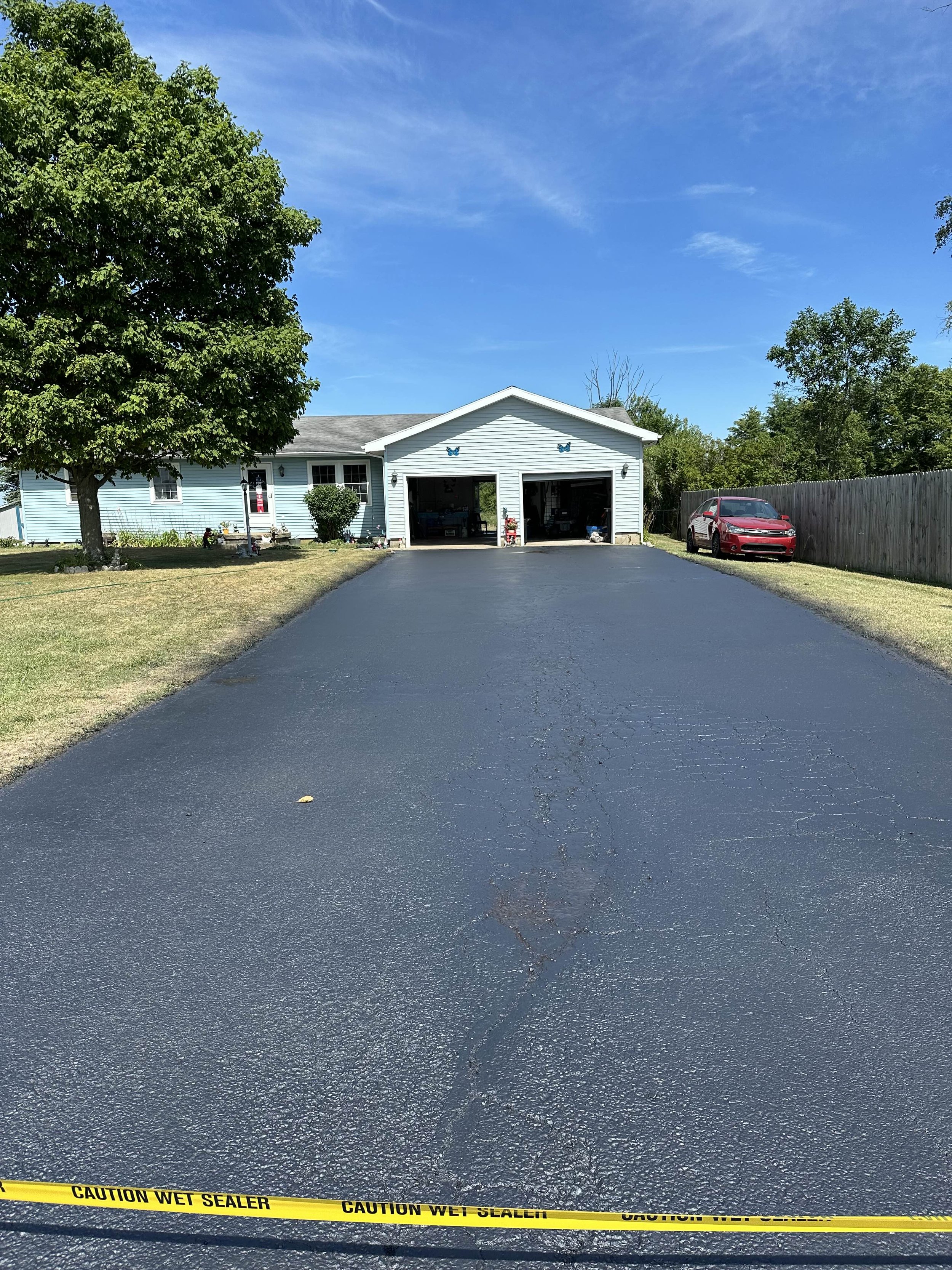 A freshly paved black asphalt driveway in front of a light blue house with white trim, surrounded by green trees and grass, with yellow caution tape reading 'Caution Wet Sealer' across the bottom.
