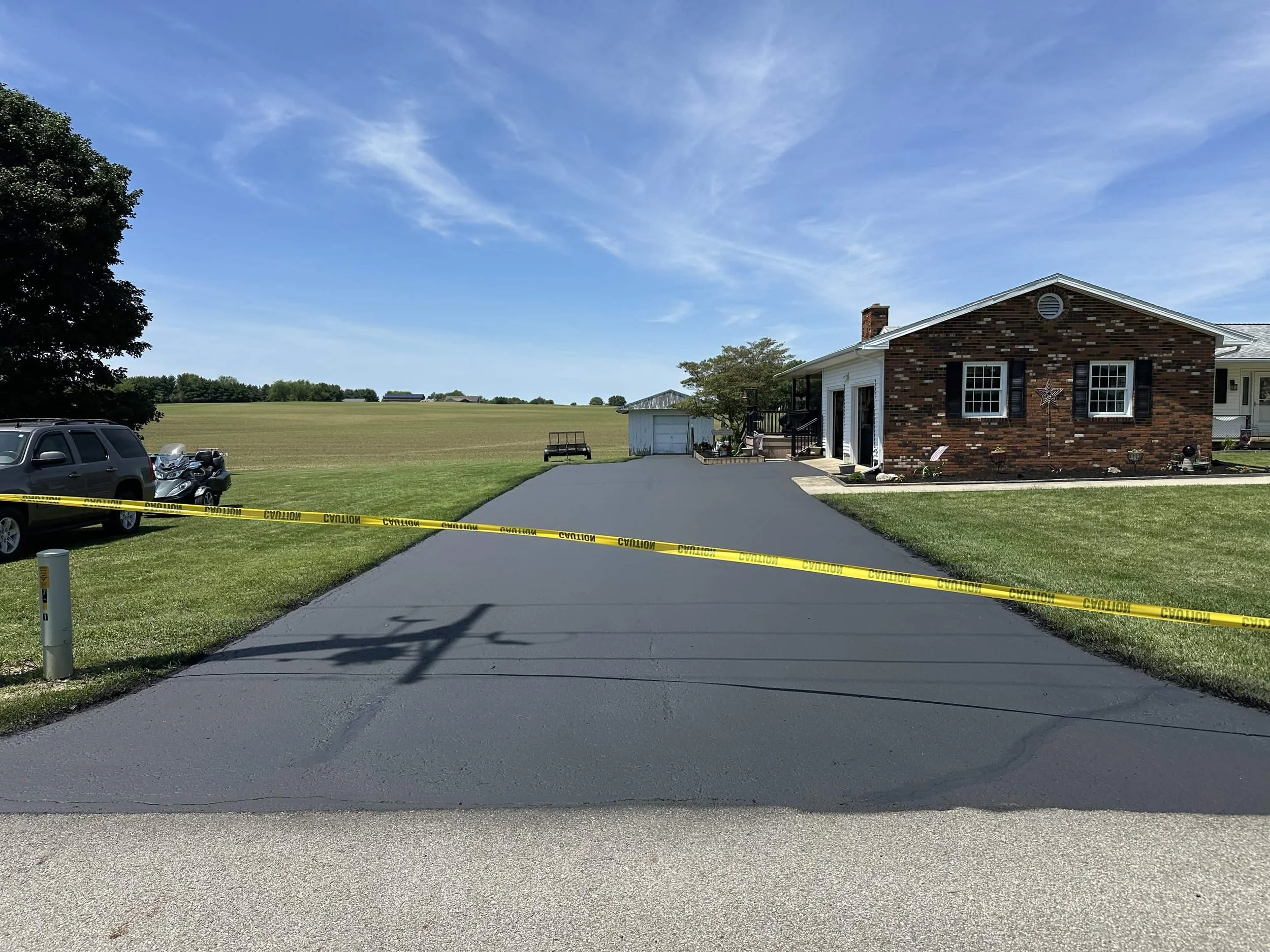 A residential driveway with caution tape across the front, leading to a brick house with a porch and black shutters, surrounded by a green yard and open field under a blue sky with clouds.