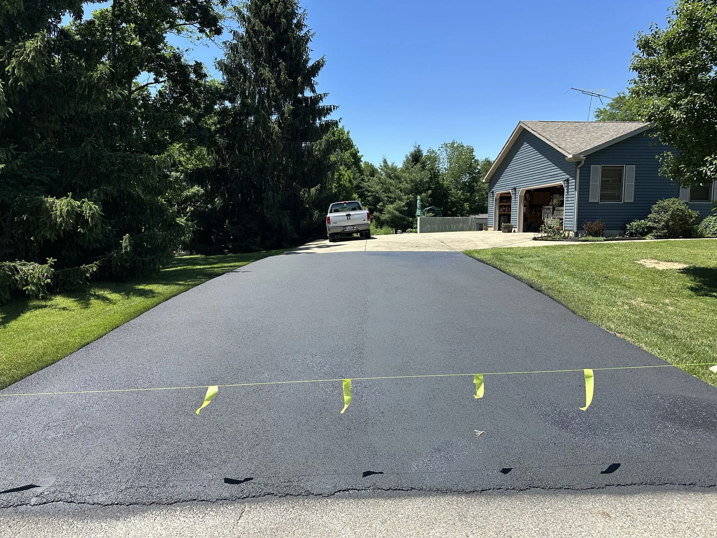 Freshly paved black asphalt driveway in front of a blue house with a garage door open, surrounded by green grass and trees under a clear blue sky.