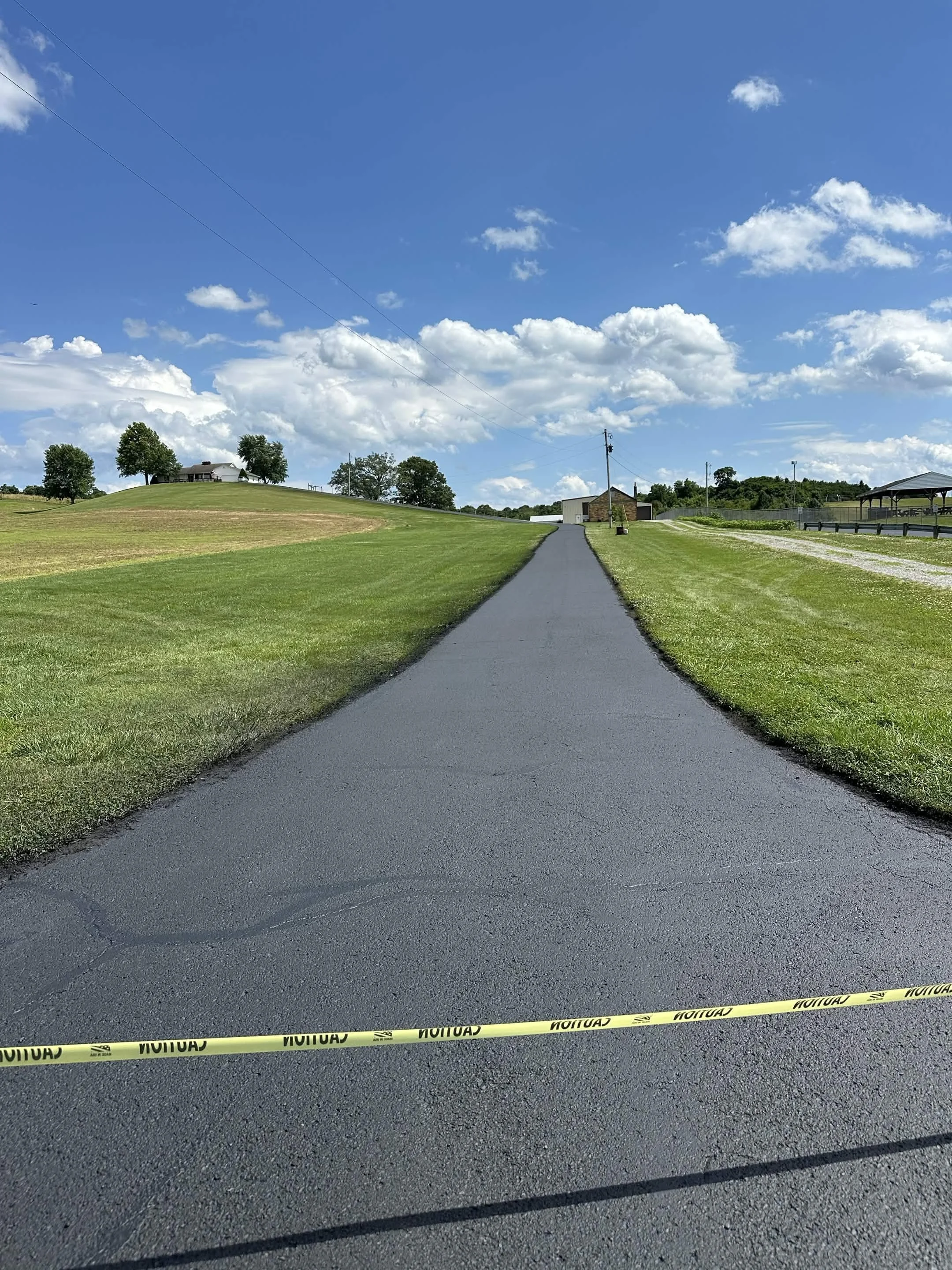 A paved road with yellow caution tape across it, situated in a grassy area with trees and buildings in the background under a partly cloudy sky.
