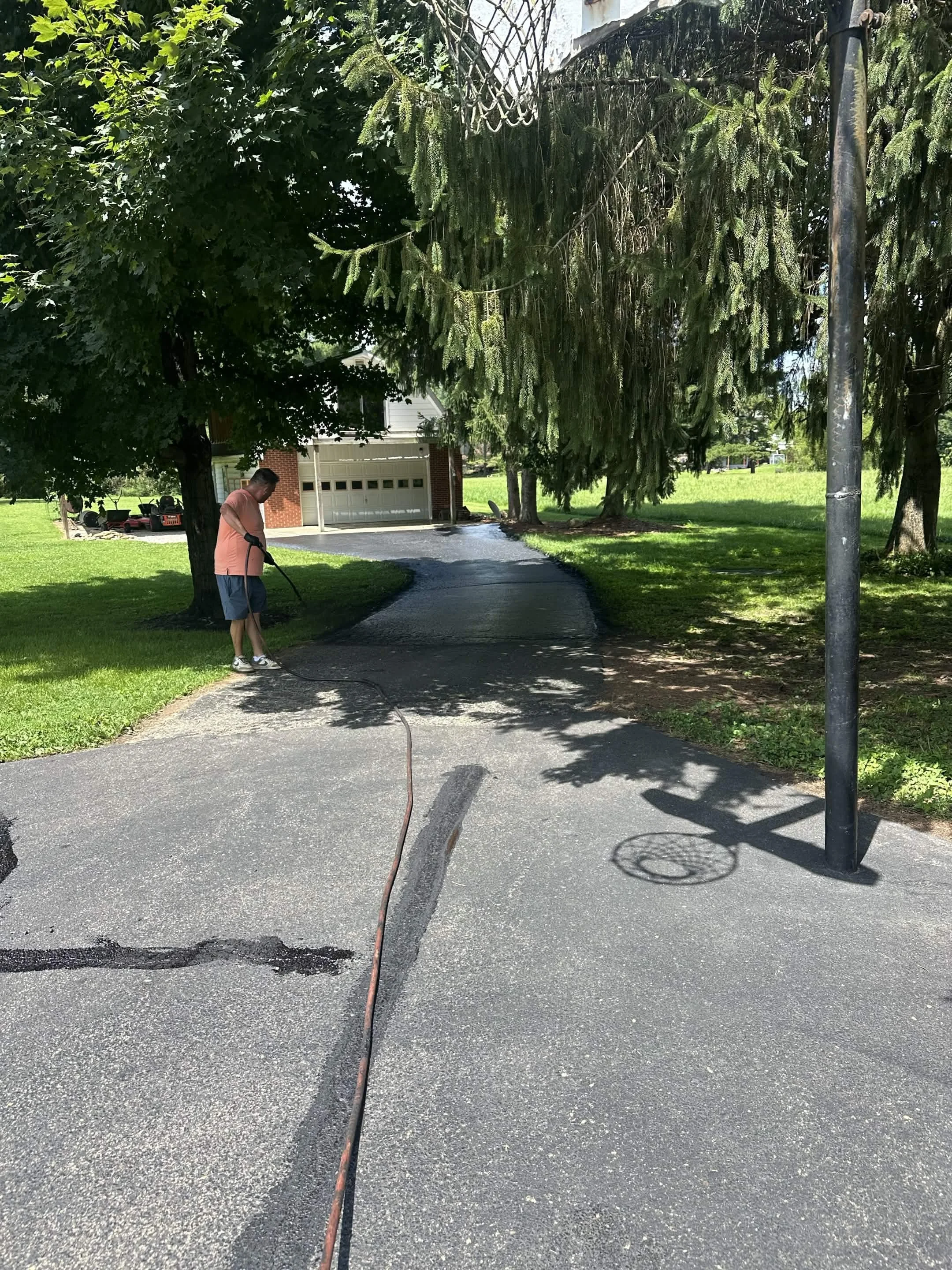A man using a pressure washer to clean the driveway of a residential house with a garage, surrounded by green trees and lawn.