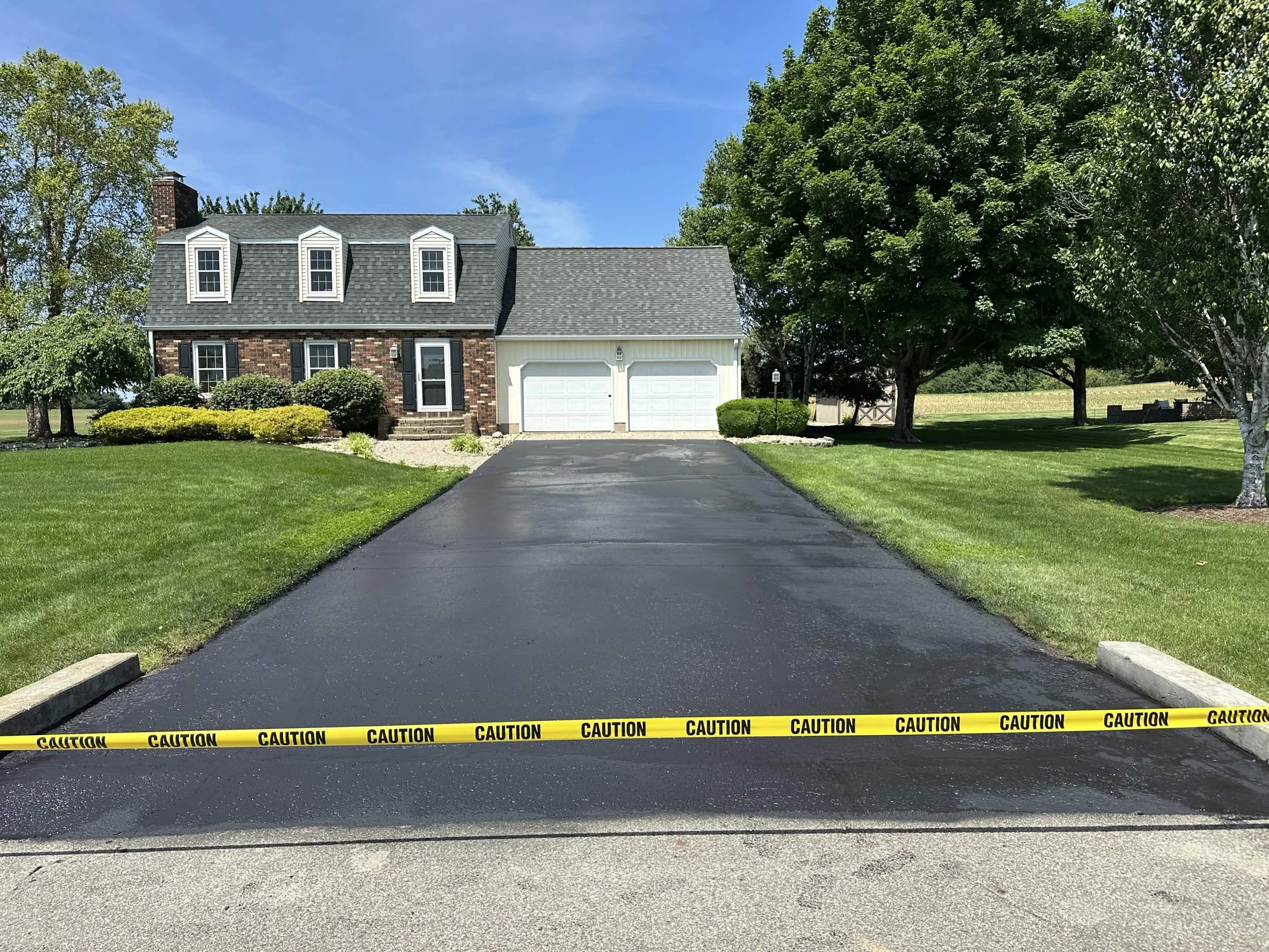 A house with a brick and white exterior, a slate roof, and a garage, surrounded by green trees and landscaped bushes, with a paved driveway and yellow caution tape across the front.