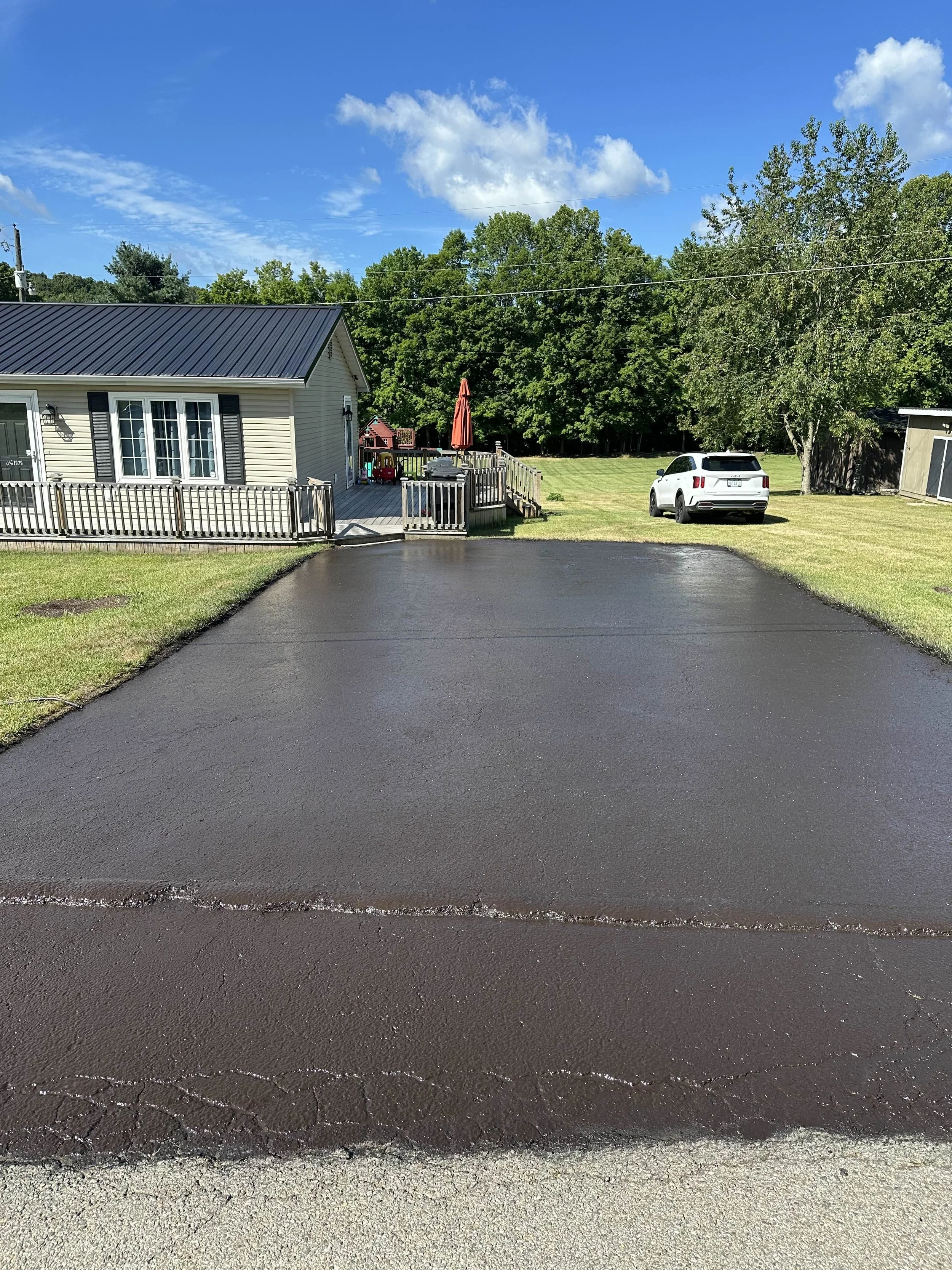 Freshly paved asphalt driveway in front of a house, with a grassy yard, trees, and a parked white SUV under a blue sky with a few clouds.