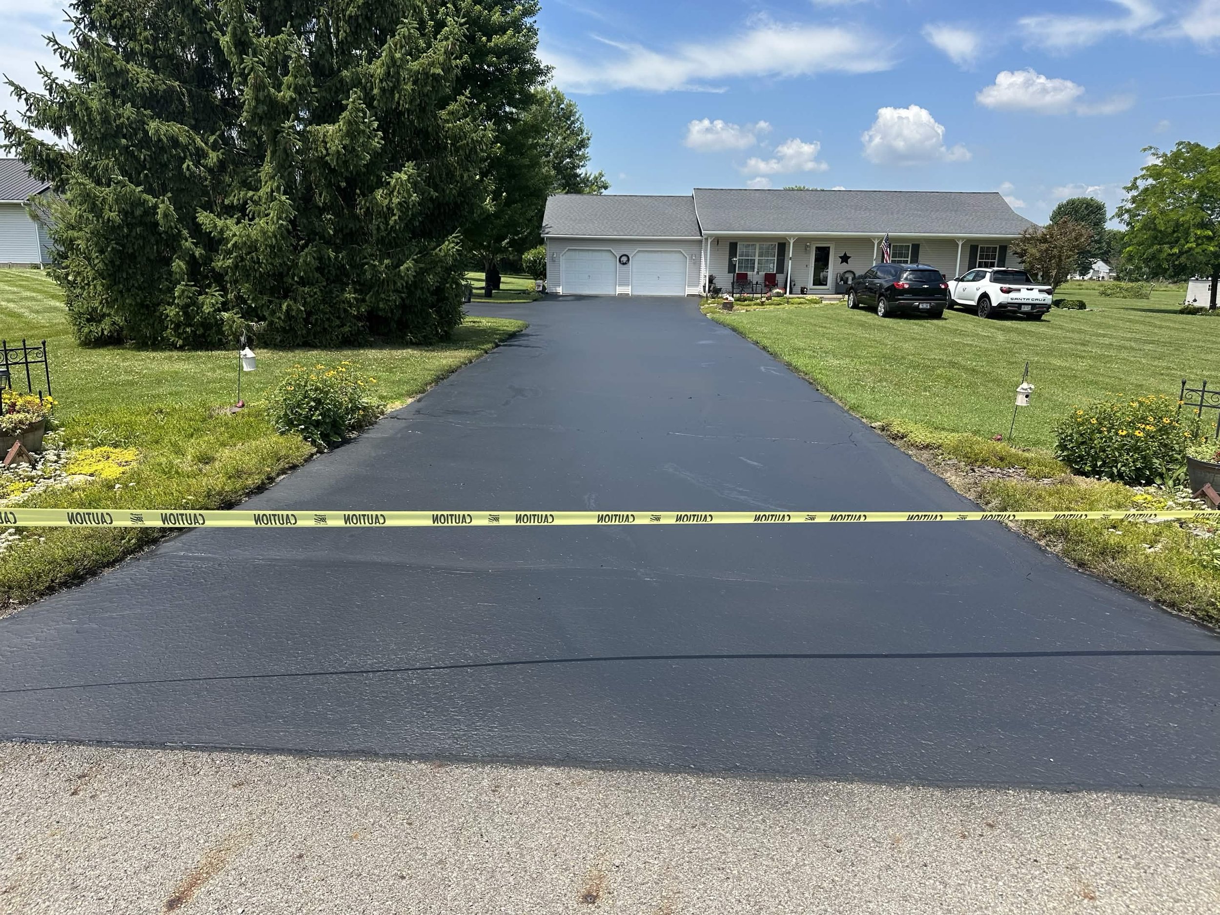 Front view of a house with a driveway, parked cars, trees, and a lawn. Caution tape is across the foreground of the driveway.