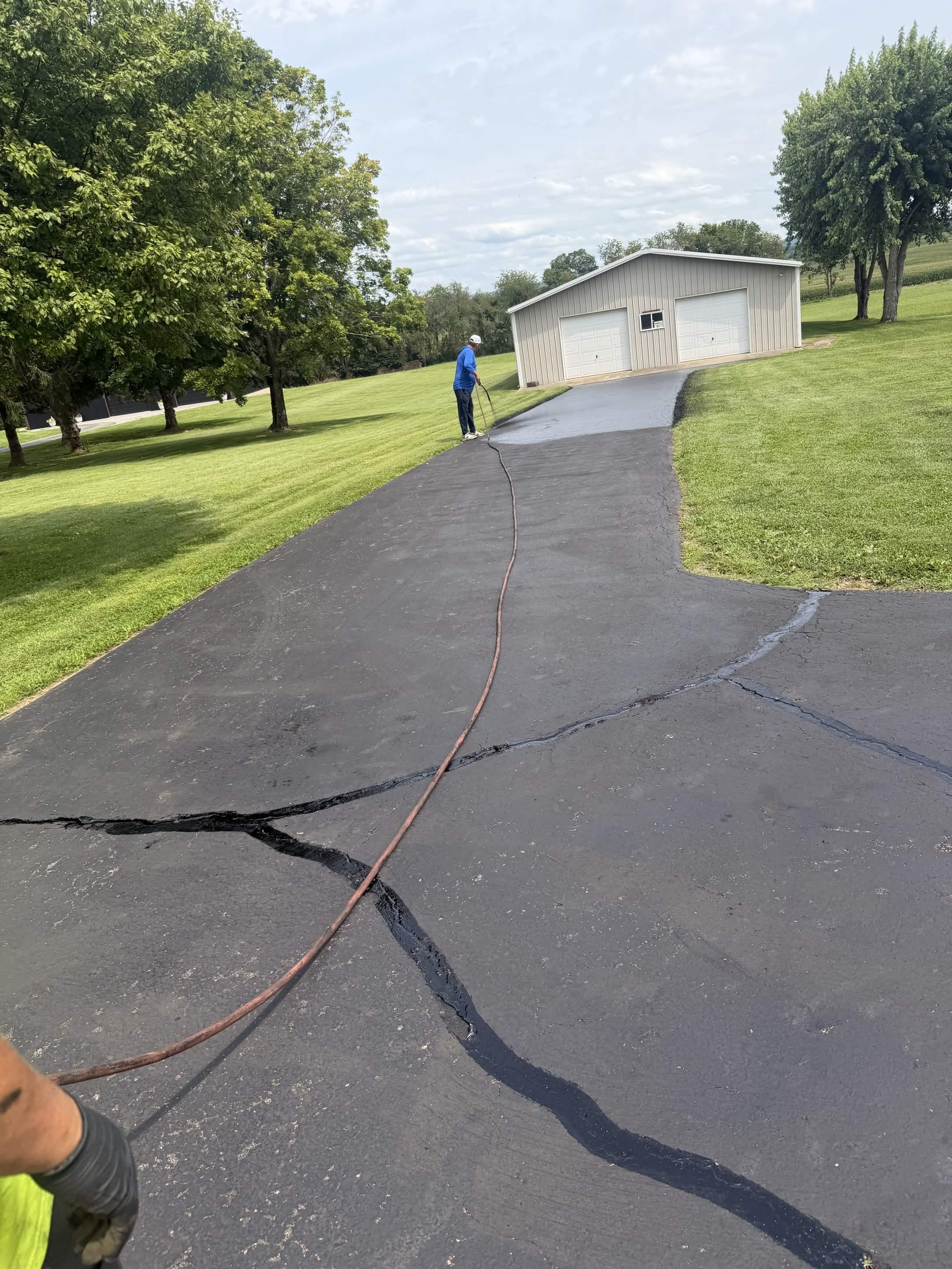 Person in blue shirt and white hat uses a pressure washer to clean a cracked asphalt driveway, with a metal garage and green trees in the background.