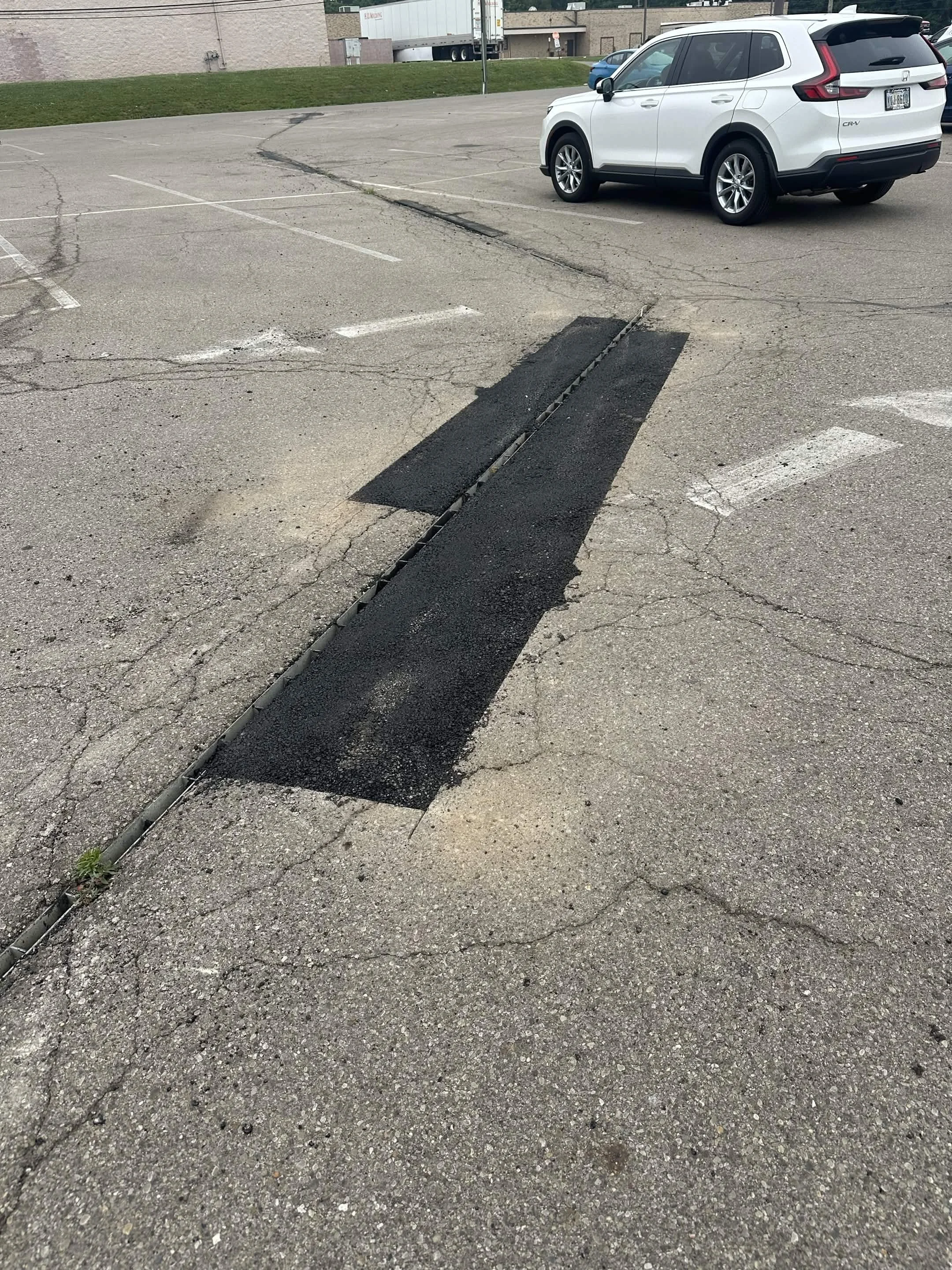 Empty parking lot with a new black asphalt section and a white SUV parked nearby. Cracks in the pavement and a small patch of grass are visible.