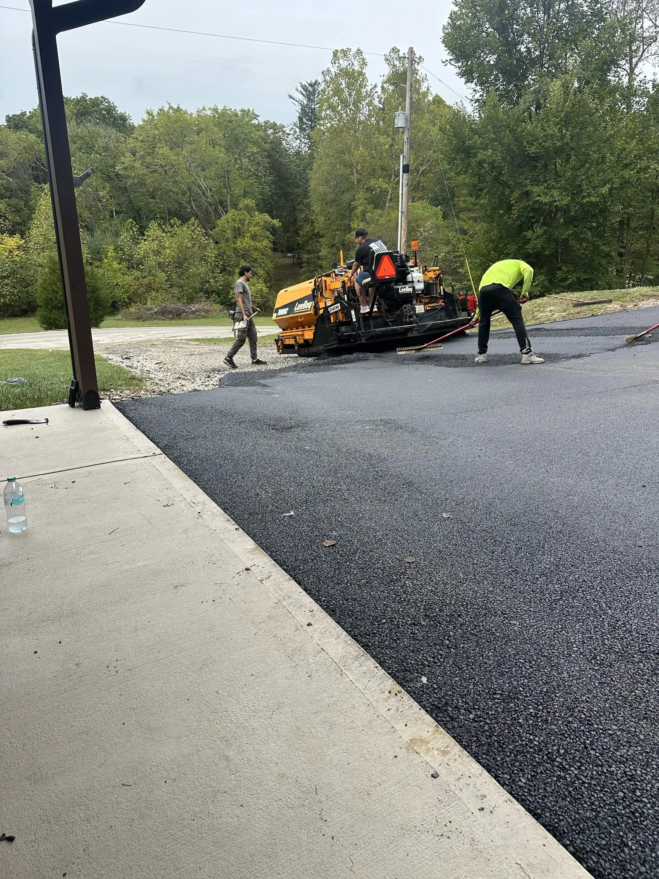 Workers paving a asphalt road next to a grassy area with trees in the background.