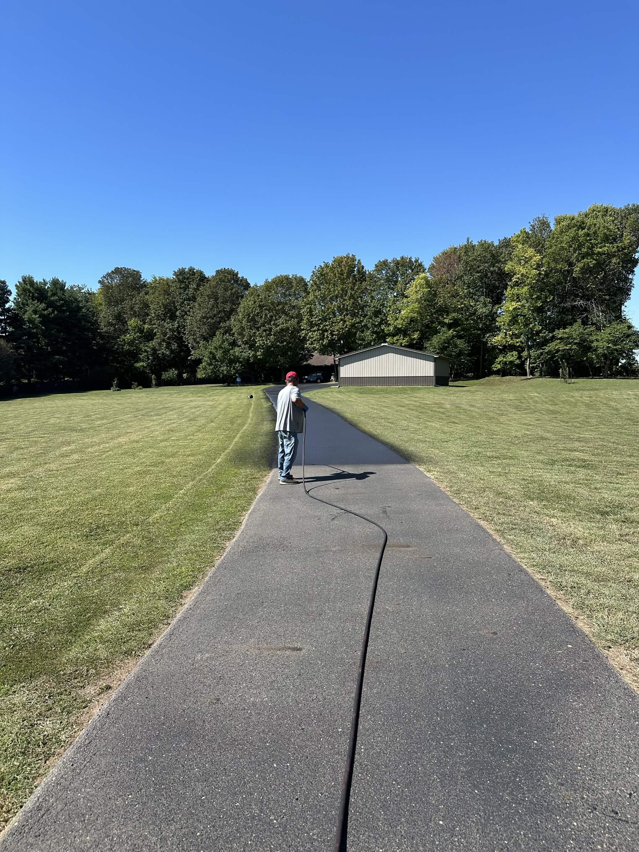 A man with a red cap and gray shirt is standing on a paved path, holding a black hose, with a grassy field, trees, and a shed in the background under a clear blue sky.