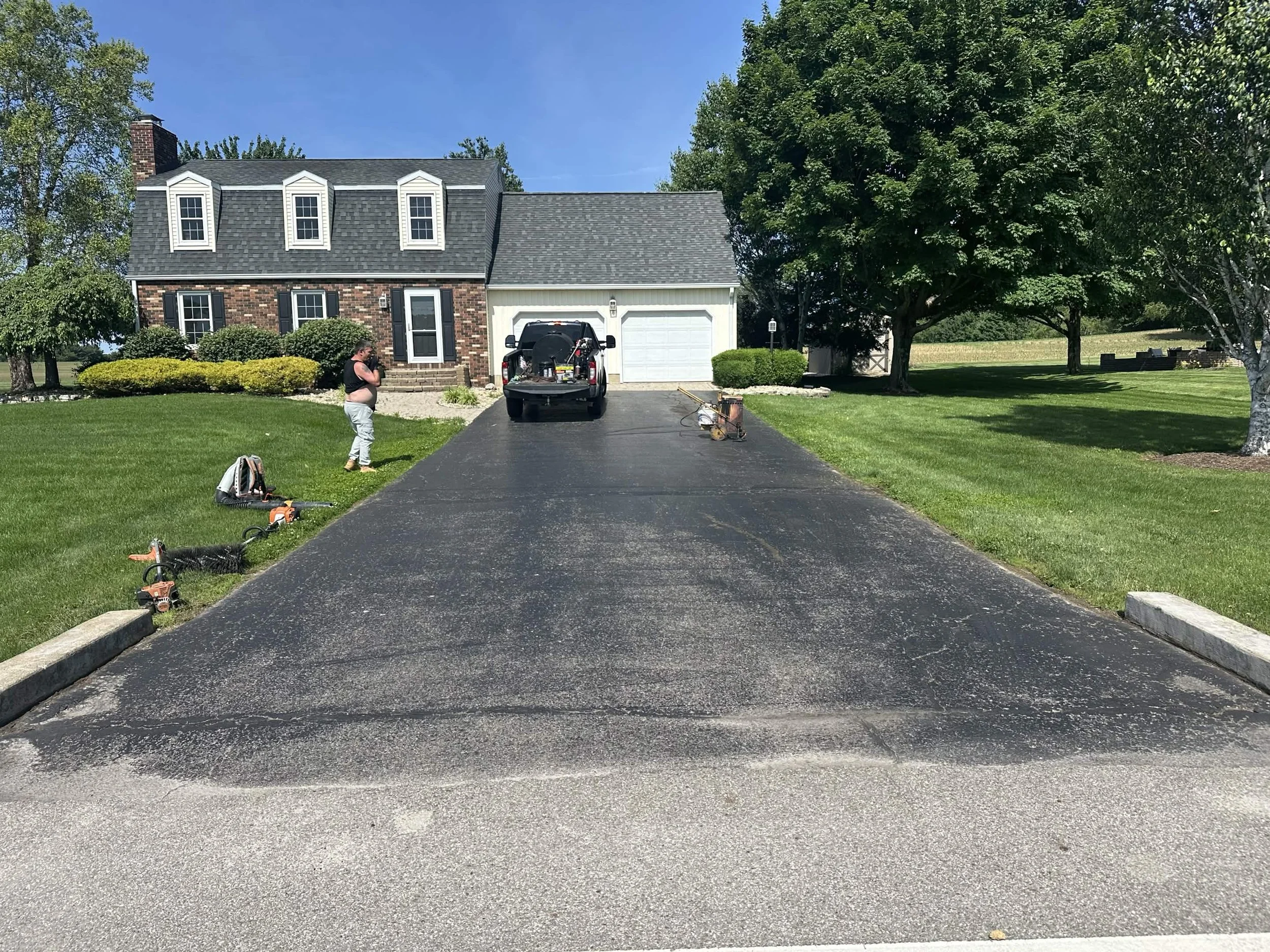 Freshly paved asphalt driveway leading to a house with a brick and siding exterior, surrounded by green grass and trees, under a clear blue sky.