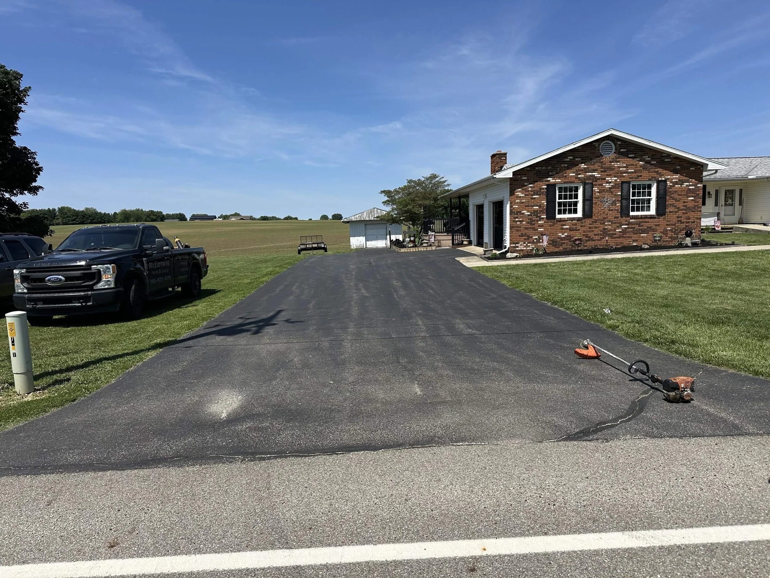 A newly paved blacktop driveway leads up to a house with a brick and white exterior. There are parked vehicles, including a black pickup truck, on the grass nearby. The sky is clear with some wispy clouds, and there is a large open field behind the h