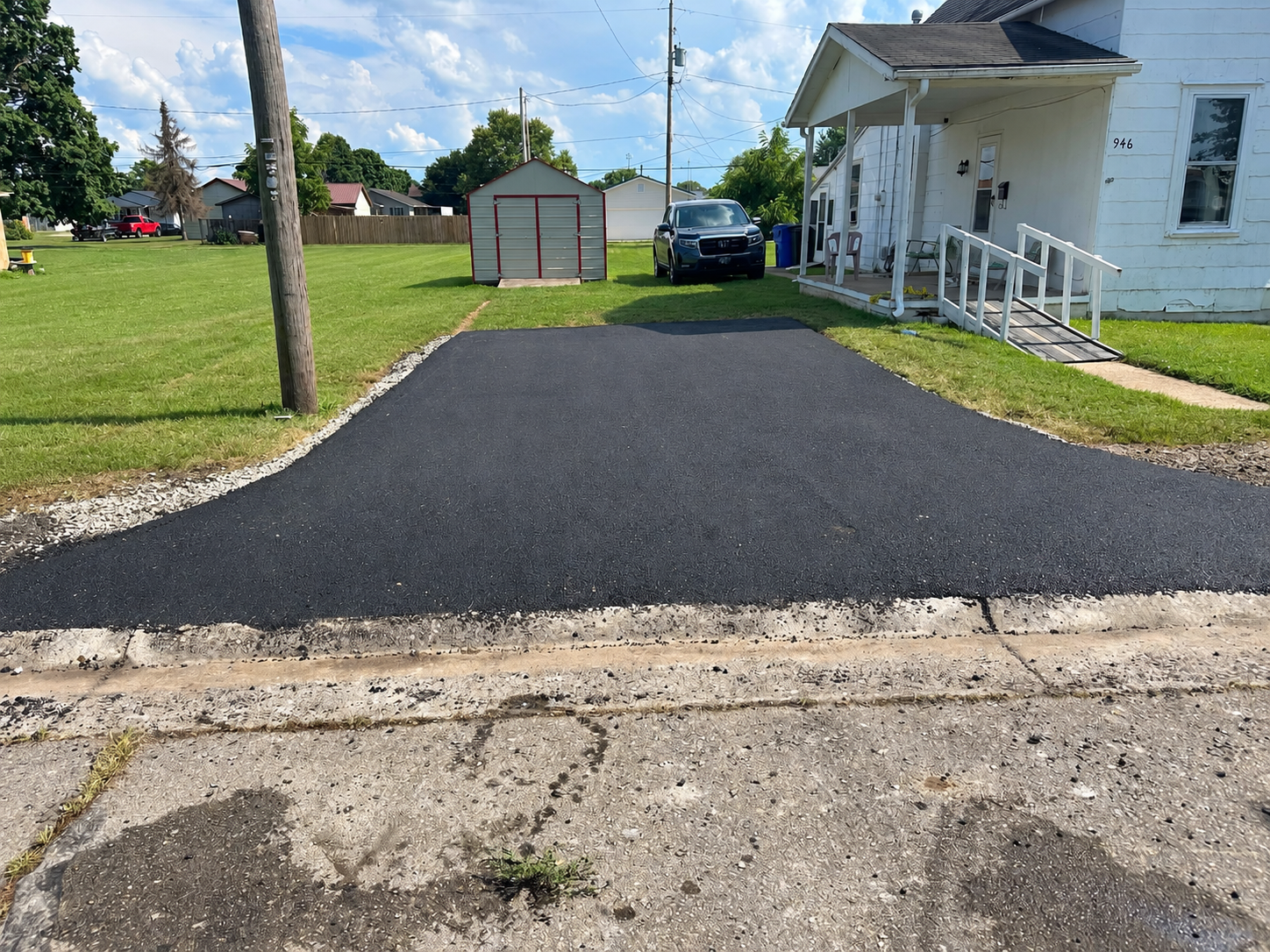 Newly paved black asphalt driveway in front of a white house with a porch and a wheelchair ramp, a black vehicle parked on the lawn, a small shed, and a swing set in a suburban yard under a clear blue sky with fluffy clouds.