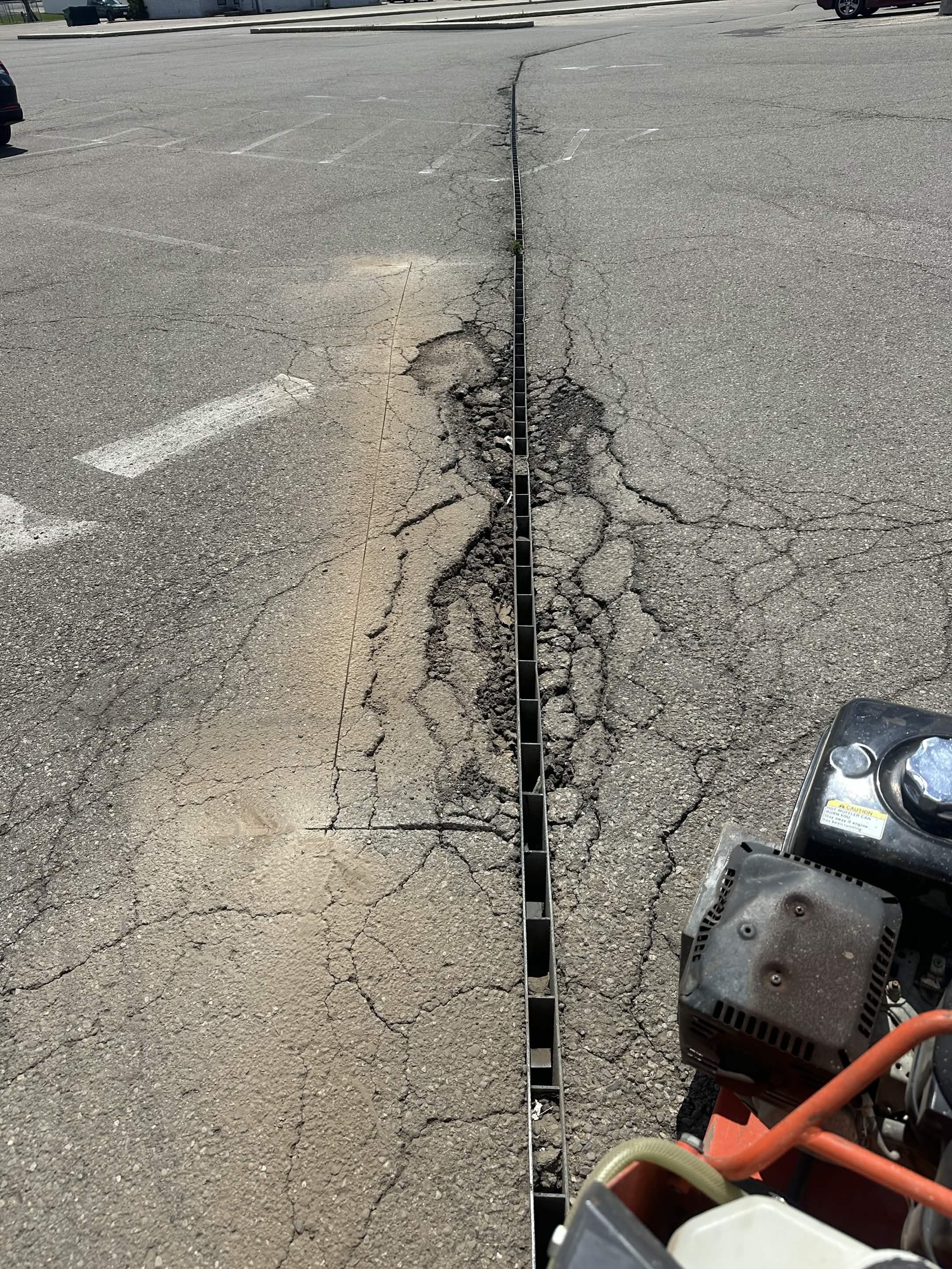 A parking lot with visible cracks and damaged pavement. A metal grate runs down the center, with some grass growing through the cracks. There are empty parking spaces and a few cars parked at the edges. Power tools are visible in the bottom right cor