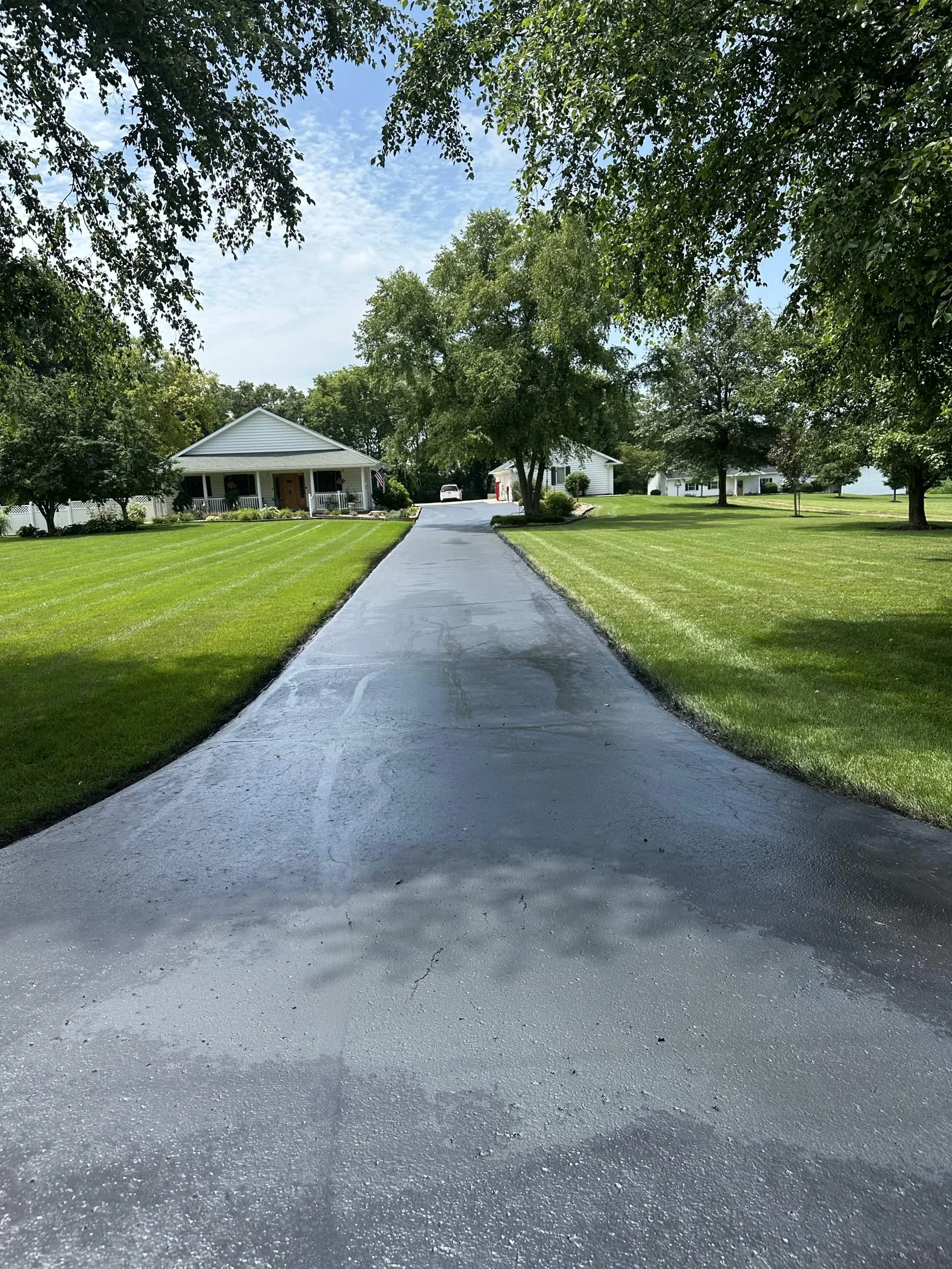 A paved driveway leads to a house with white siding, surrounded by green lawn and trees under a partly cloudy sky.