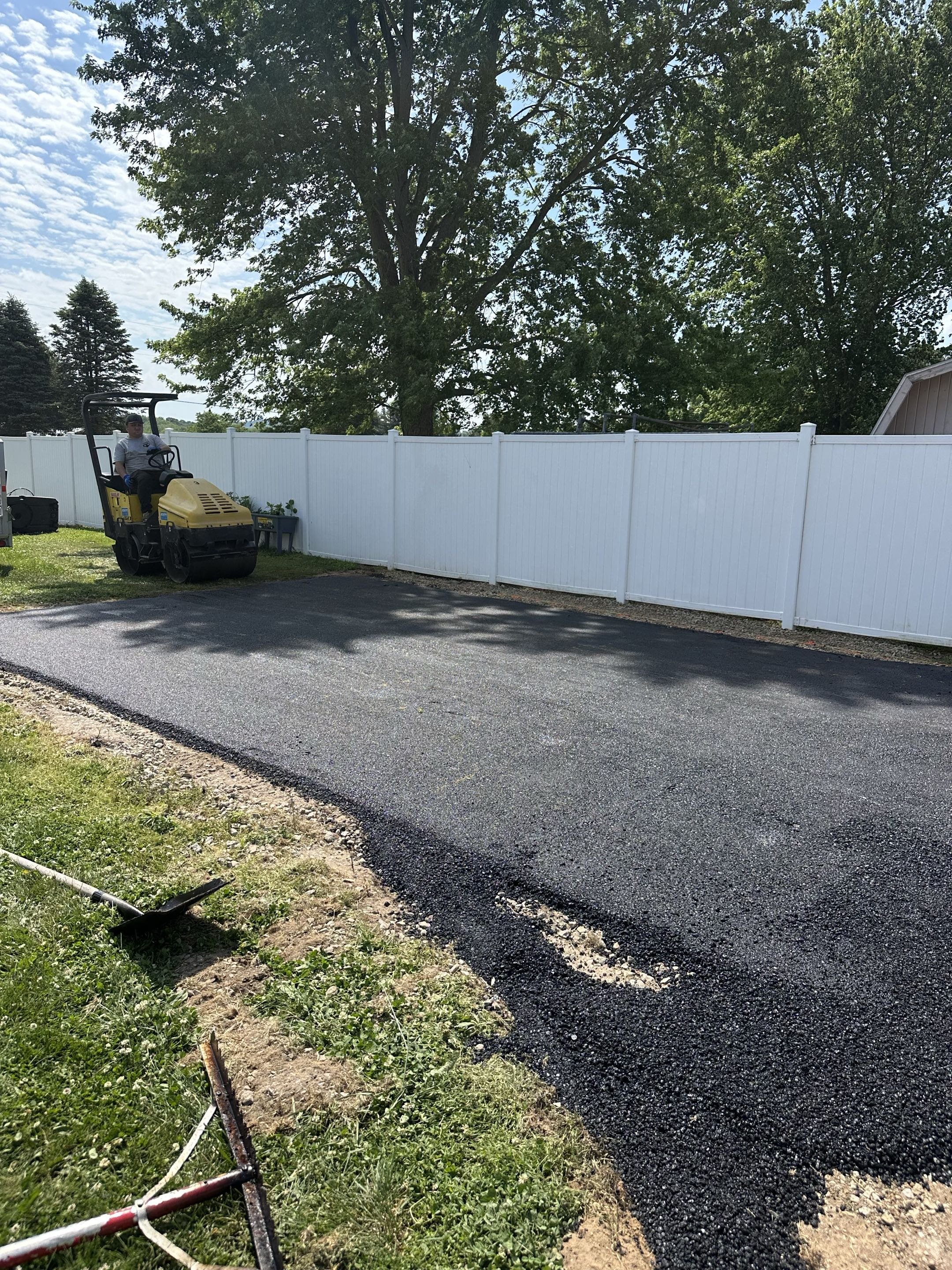 A worker on a yellow compacting machine working on a freshly laid asphalt road surface in a backyard, with a white fence, green trees, and a partly cloudy sky in the background.