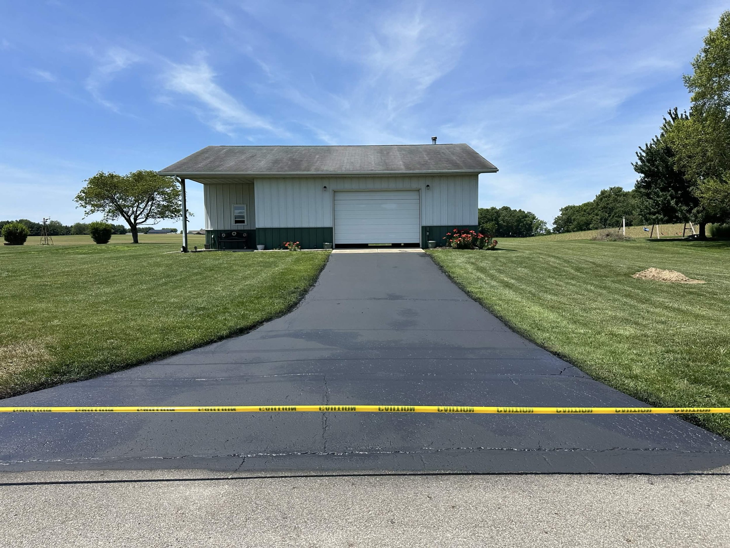 A paved driveway leading to a white shed with a closed garage door, surrounded by green grass and trees, with caution tape across the front of the driveway.
