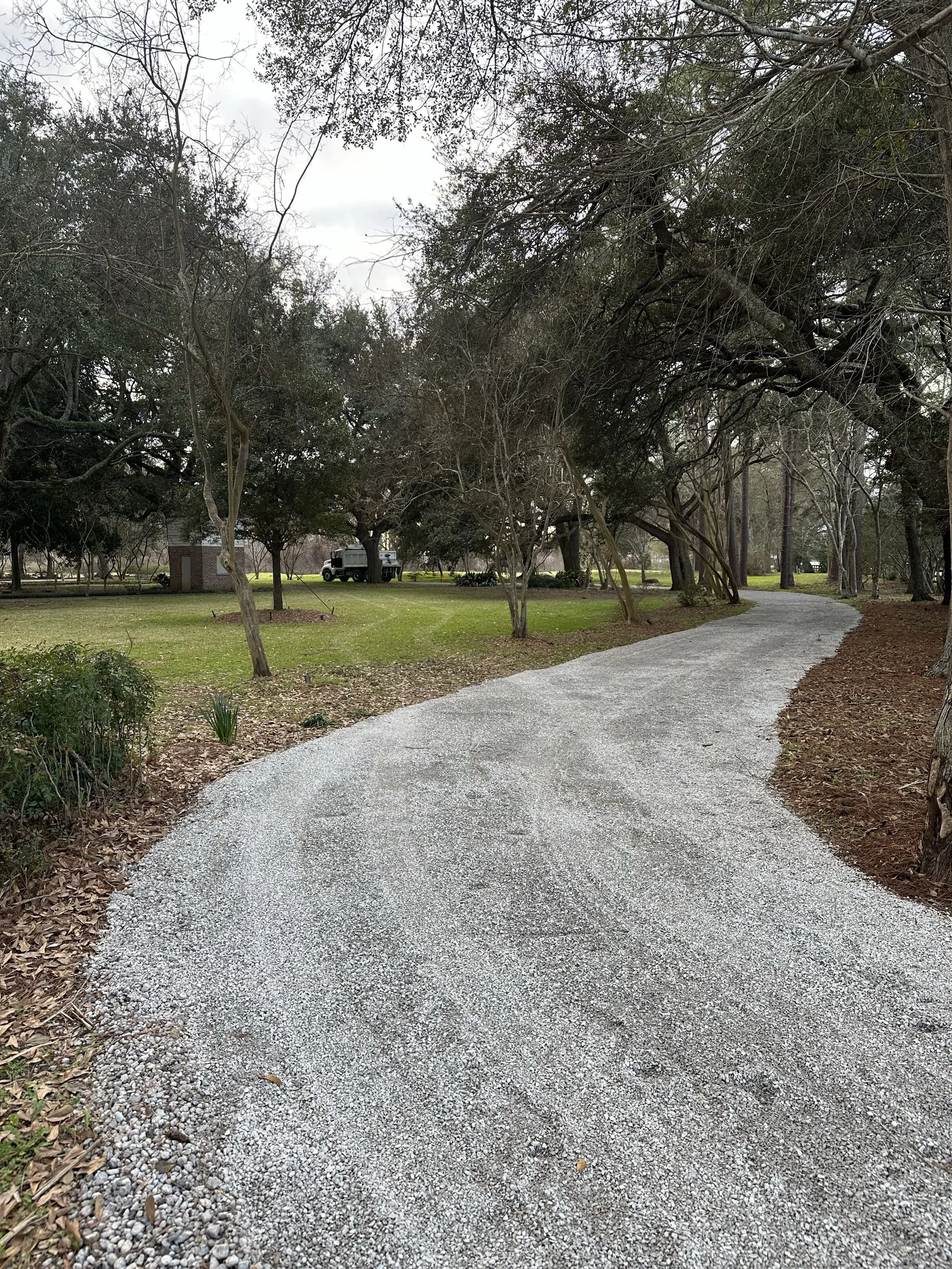 A gravel walking trail winding through a park with trees on either side, some without leaves, and a cloudy sky overhead.
