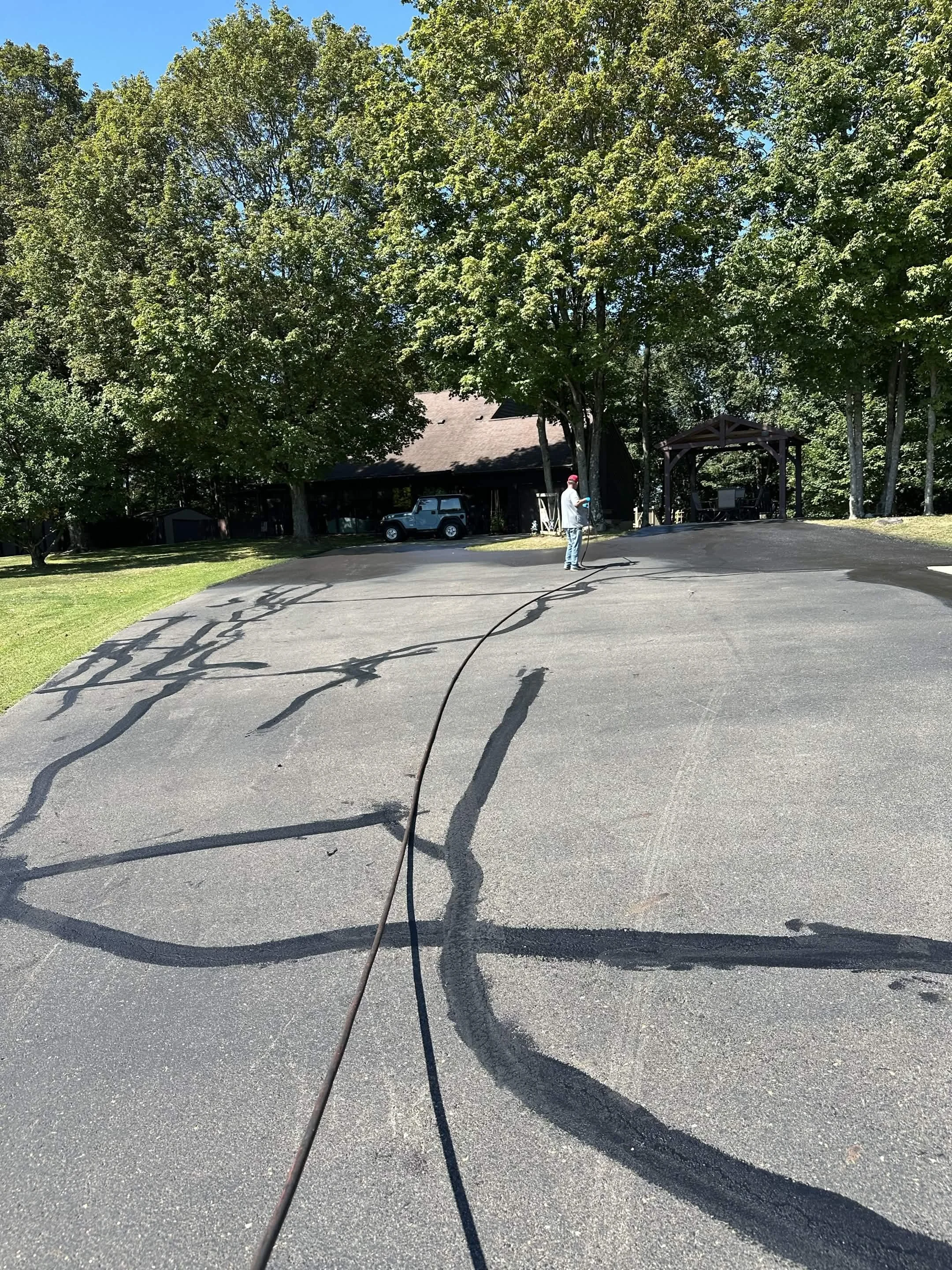 A man in a gray shirt and jeans is using a power washer on a paved driveway. There are large green trees, a house, and a parked vehicle in the background.